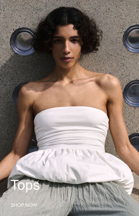 Person in a white strapless top and gray pants sits against a textured wall adorned with circular patterns. Text reads: "Tops SHOP NOW".