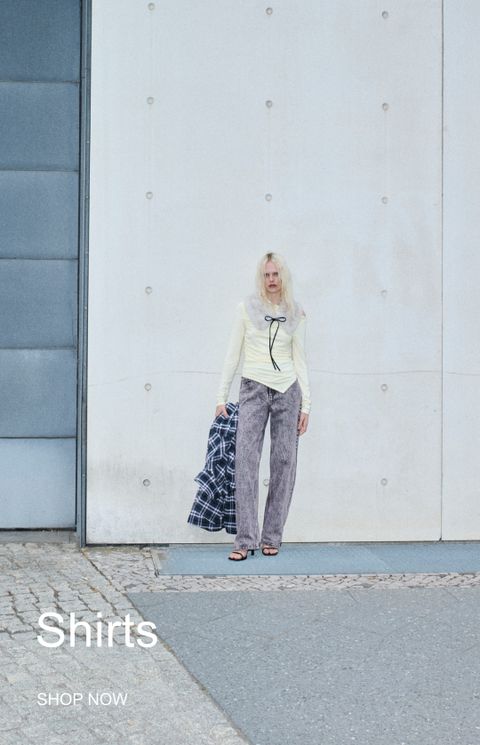 Person in a light shirt with a fur collar and gray pants, holding a checkered shirt, stands against a concrete wall. Text reads "Shirts, Shop Now."
