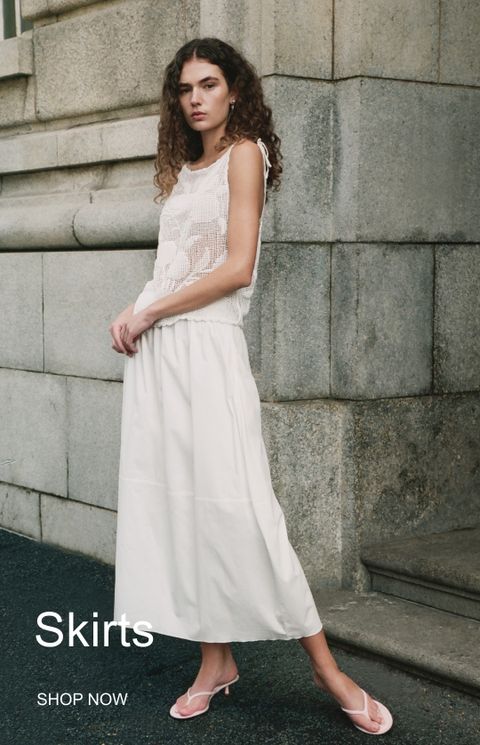 Woman in a white crocheted top and long skirt stands against a stone wall, wearing sandals. Text reads "Skirts" and "Shop Now."