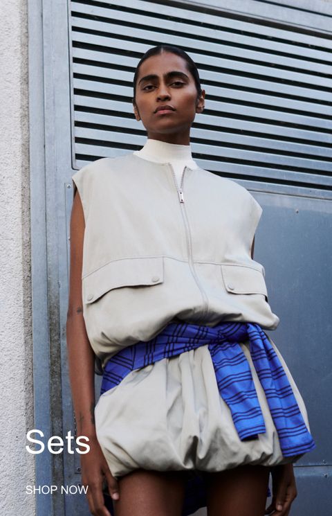 Person in a sleeveless beige top with a blue plaid shirt tied at the waist, standing against a metal louvered background. Text: "Sets, SHOP NOW".