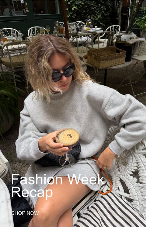 Woman in sunglasses sips a cocktail at an outdoor cafe. She's wearing a gray sweatshirt. Text reads "Fashion Week Recap - Shop Now."