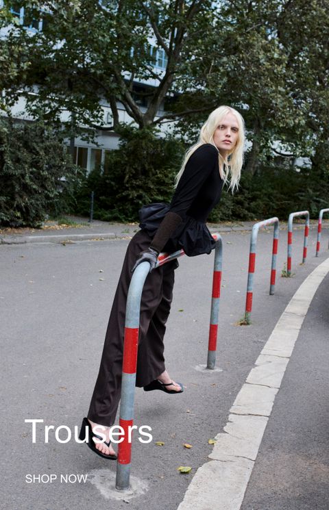 A person in a stylish black top and dark trousers leans on a red and gray pole in an urban setting, with "Trousers" text overlay.
