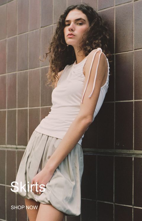 A woman with curly hair wearing a white top and beige skirt leans against a tiled wall. Text reads "Skirts" and "Shop Now" in the corner.