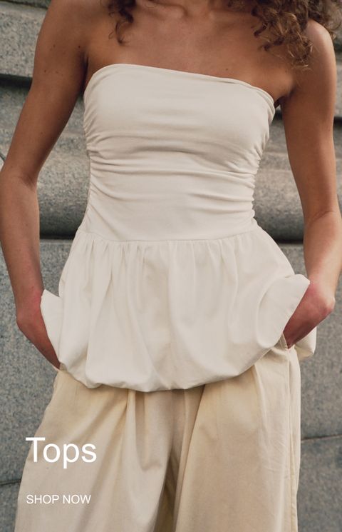 A woman wearing a strapless white top with a gathered hem and tan pants, standing in front of a stone wall. Text reads "Tops, Shop Now."