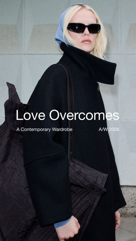 Model wearing a black coat and sunglasses, with a light blue hood, carrying a large, textured dark tote bag. Text reads "Love Overcomes" and A/W 2026.