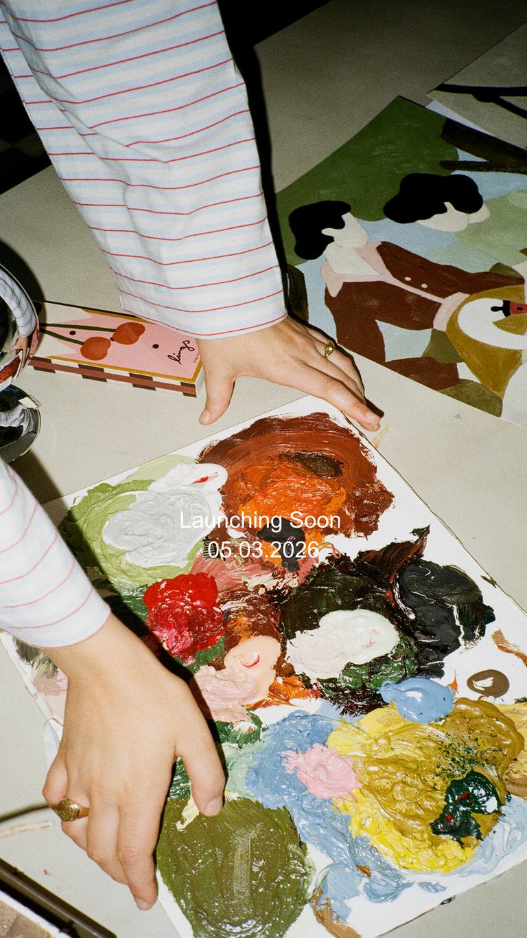 Hands with striped sleeves holding abstract artwork with various paint colors. Launch date text and paint palette nearby on a table.