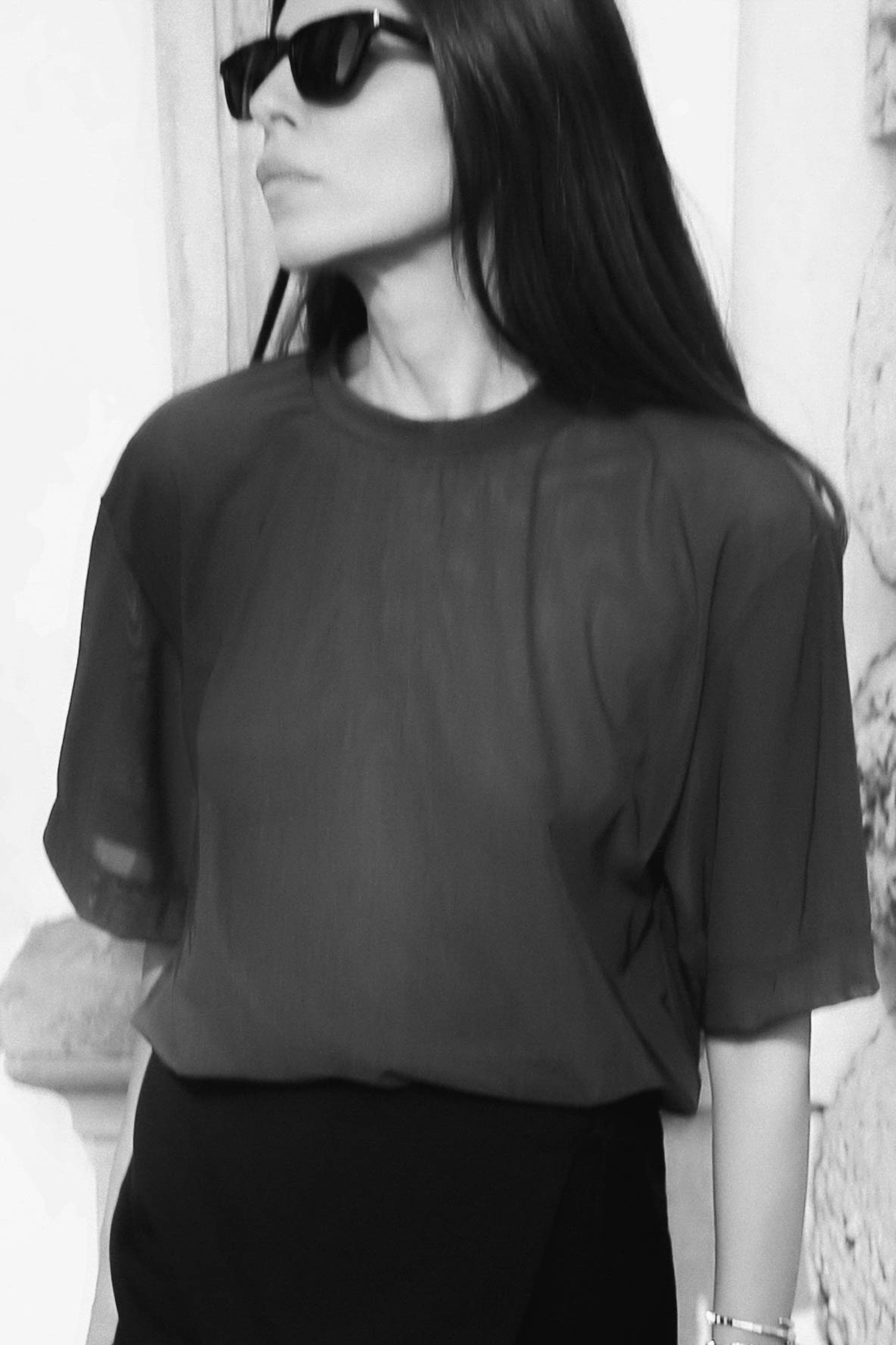 Woman in sunglasses wearing a sheer blouse, looking to the side, in black and white.