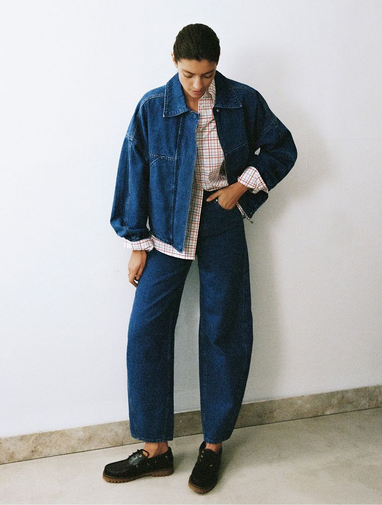 Person wearing a denim jacket, plaid shirt, and blue jeans, standing against a white wall, looking down with one hand in pocket.