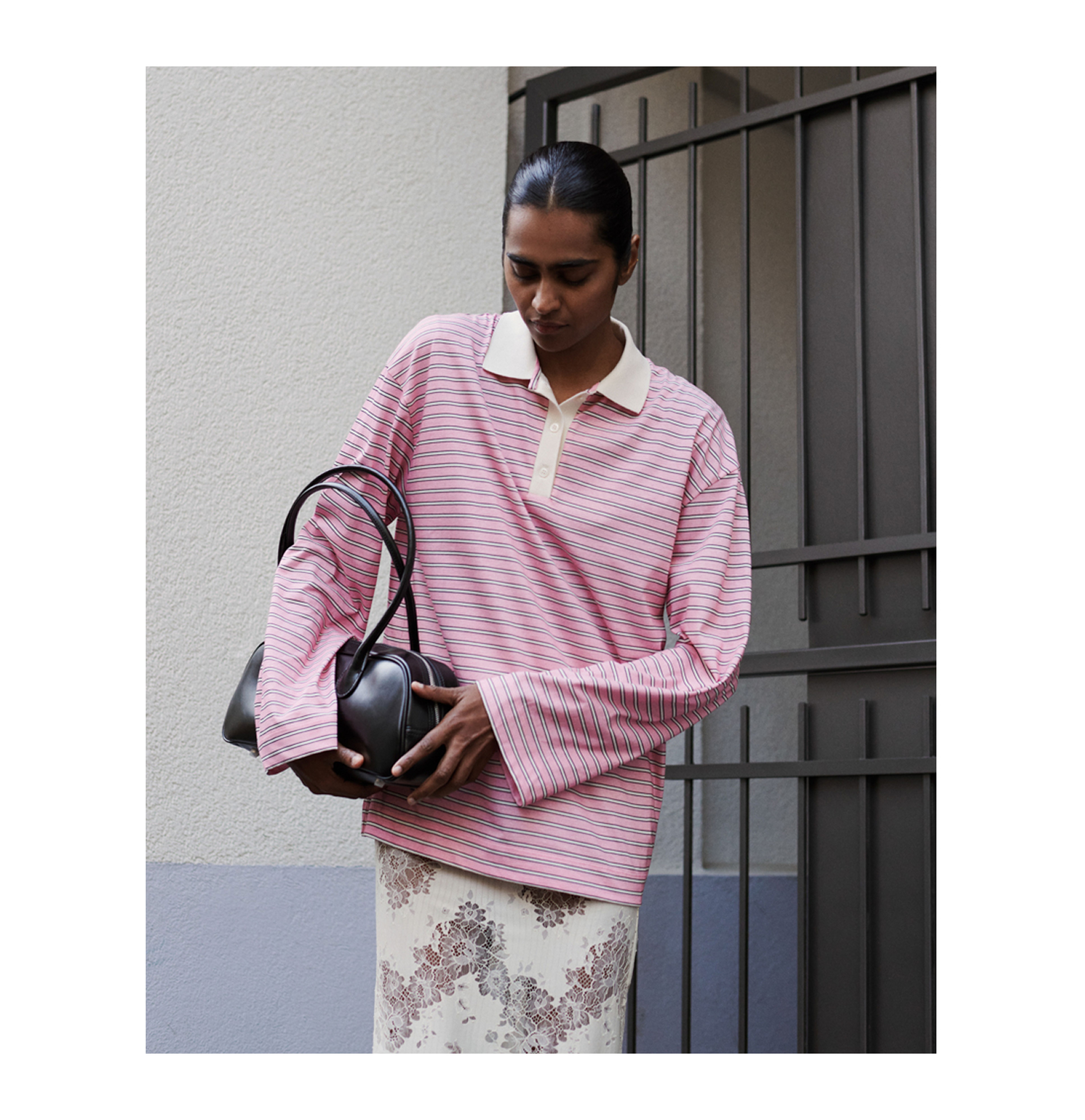 Person in a pink striped shirt and floral skirt holds a black bag, standing by a metal gate against a light wall.