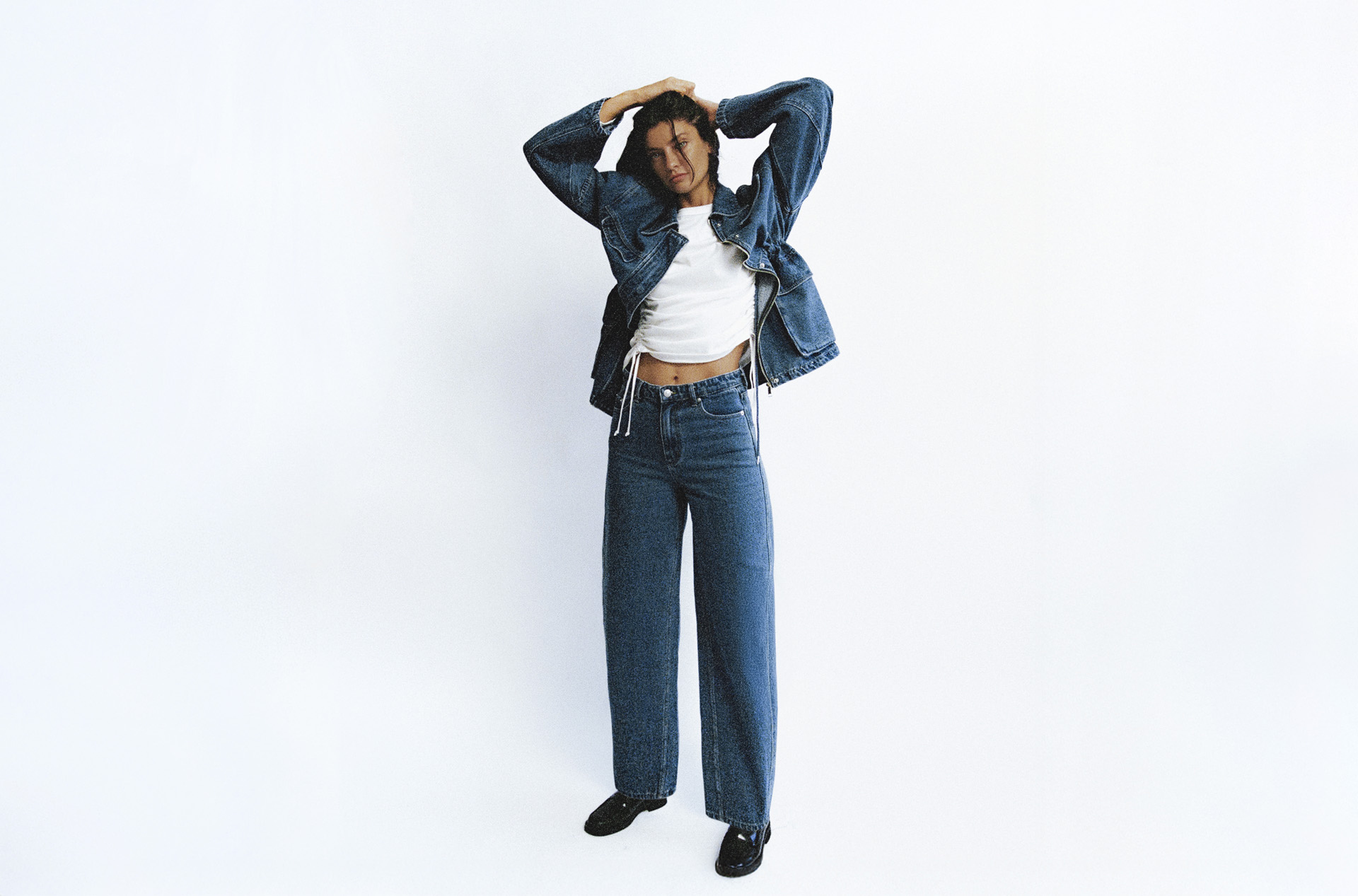 Person posing in a white shirt, denim jacket, and wide-leg jeans, with hands resting on their head against a plain white background.