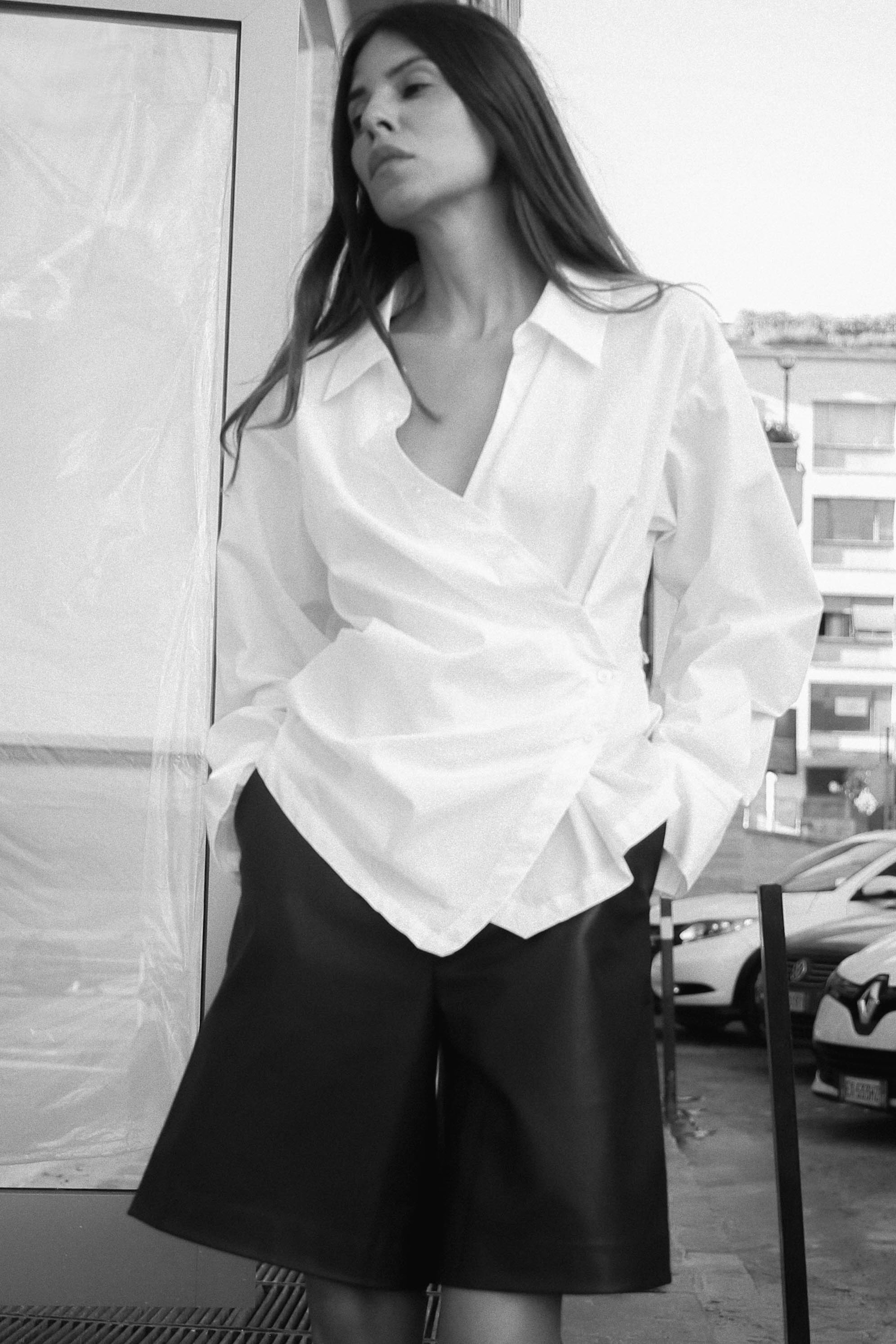 Woman in a white wrap blouse and black shorts leans against a wall, looking away, with a blurred urban background. Black and white photo.