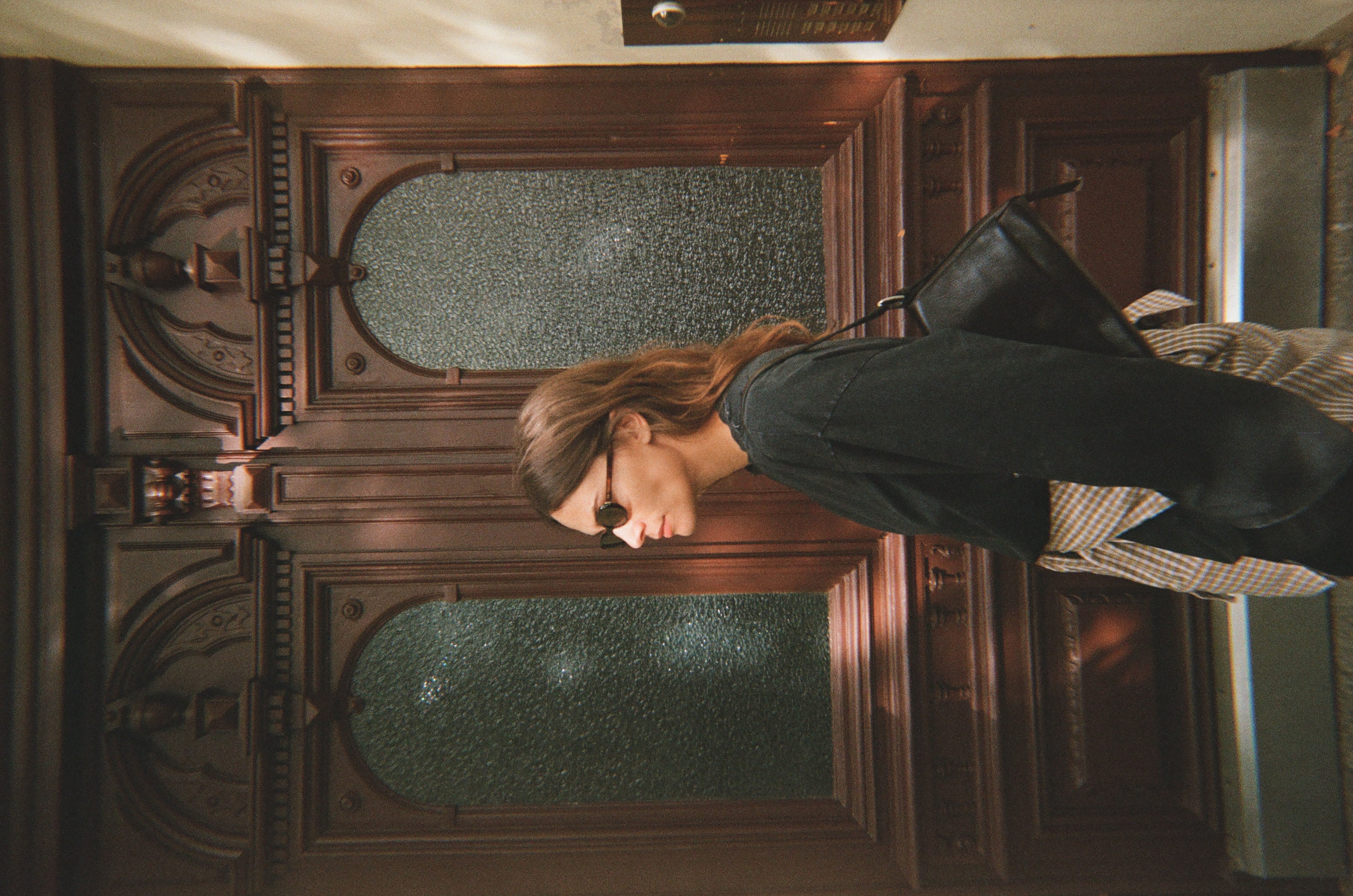 Woman in sunglasses and dark clothing stands in profile in front of a decorative wooden door.