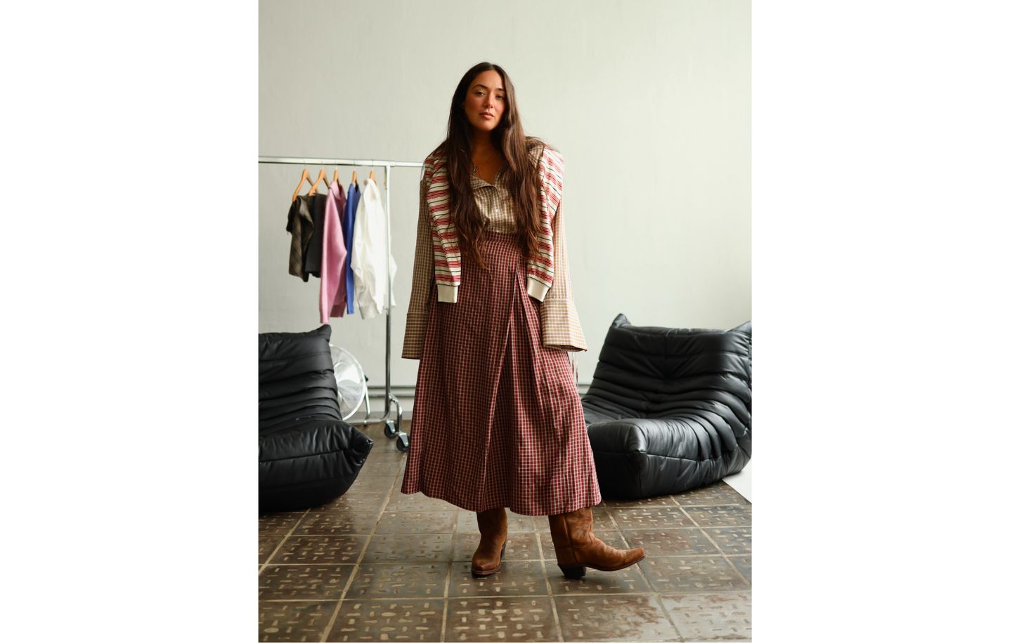 Woman in a long plaid skirt and cowboy boots poses in a room with clothes rack and black lounge chairs.