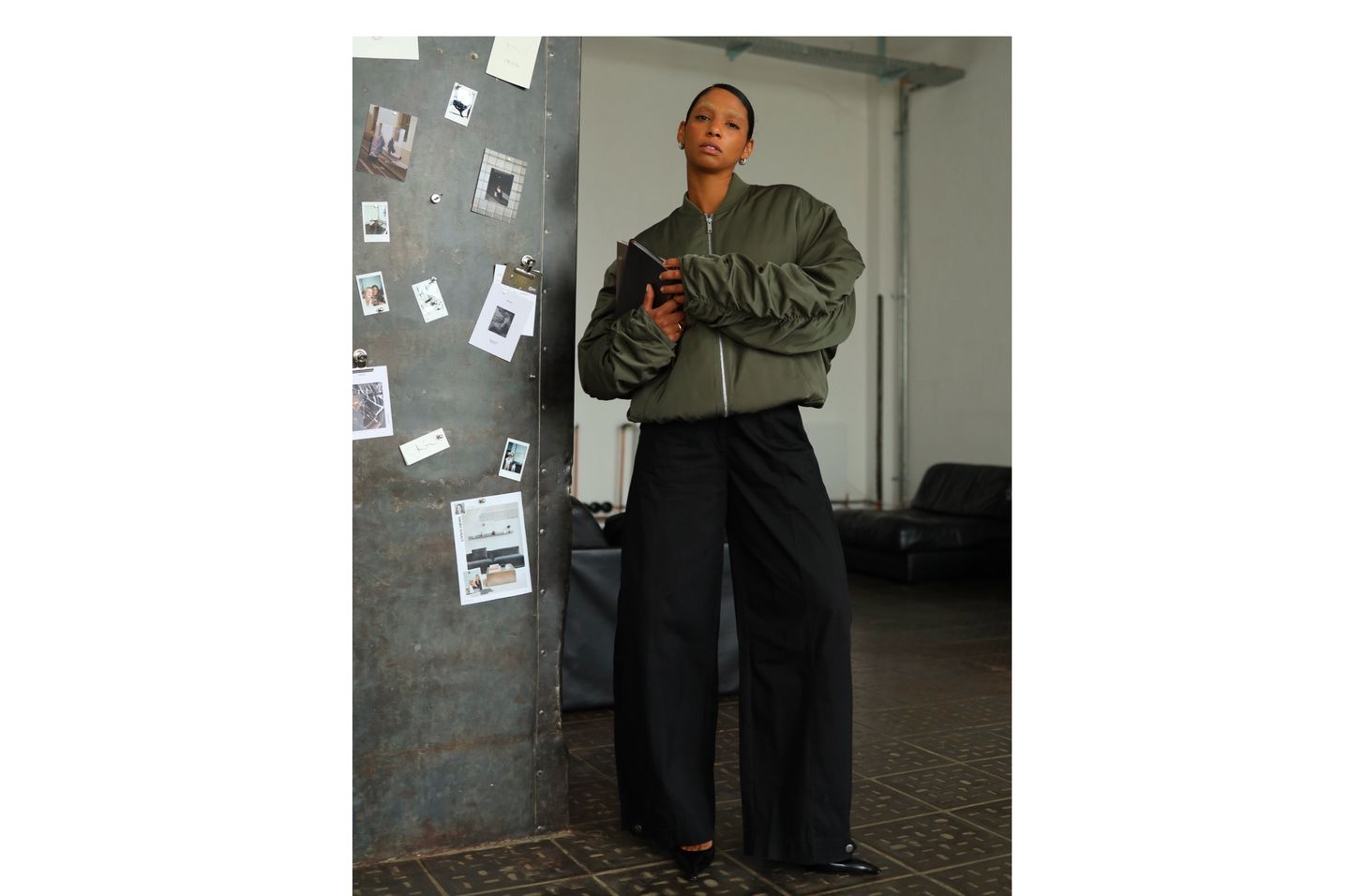 Person in an oversized olive bomber jacket and black wide-leg pants stands by a bulletin board in a minimalist room.