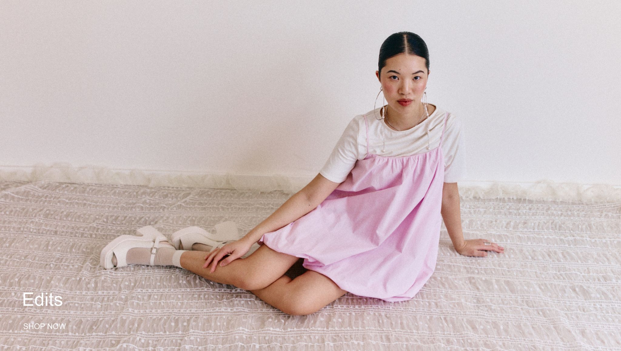 Woman in a pink dress sits gracefully on a patterned floor, styled with white shoes and a white shirt, against a plain backdrop.