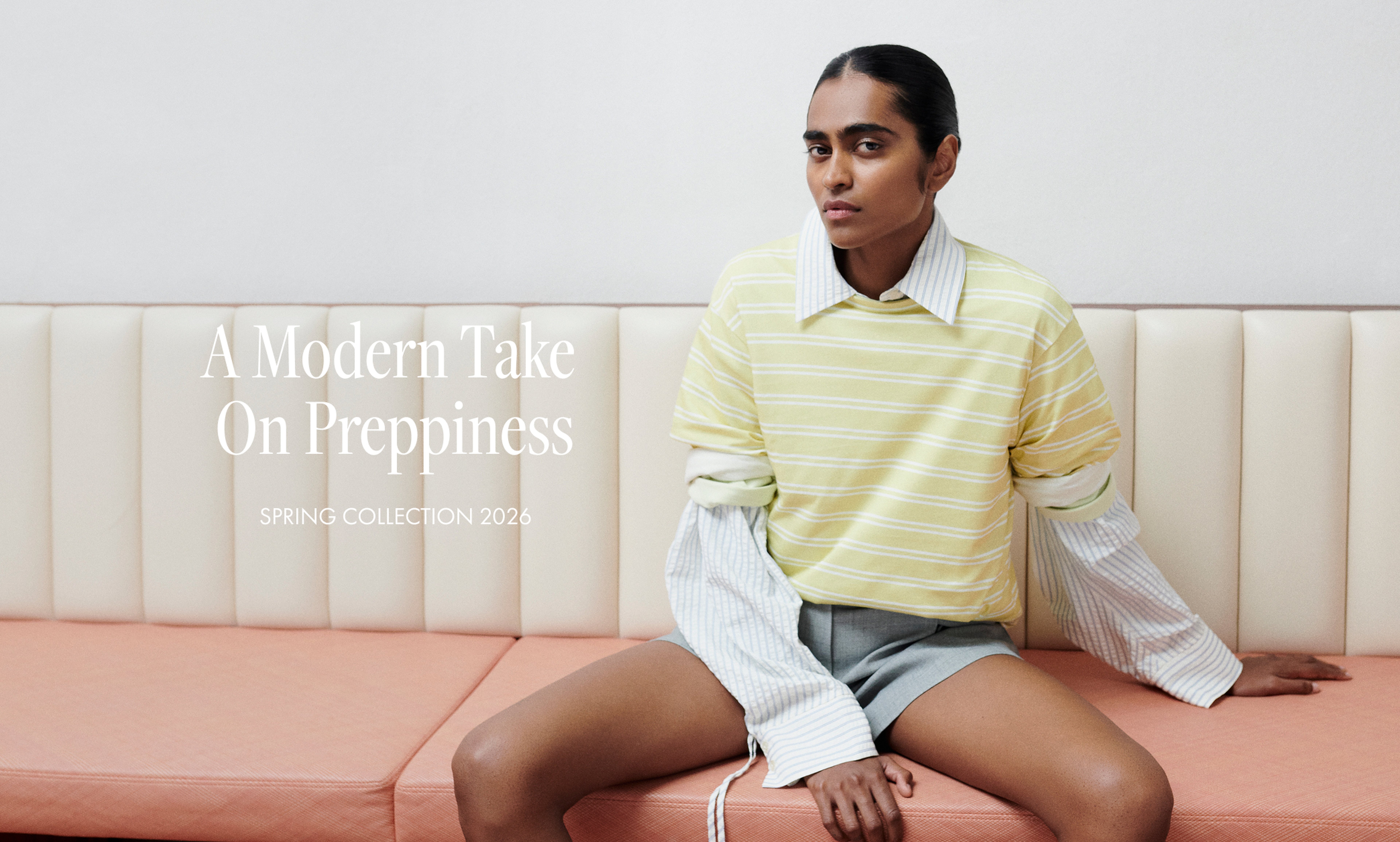 Person sitting on a sofa wearing a striped yellow shirt over a long-sleeve, showcasing a modern, preppy style from the Spring 2026 collection.
