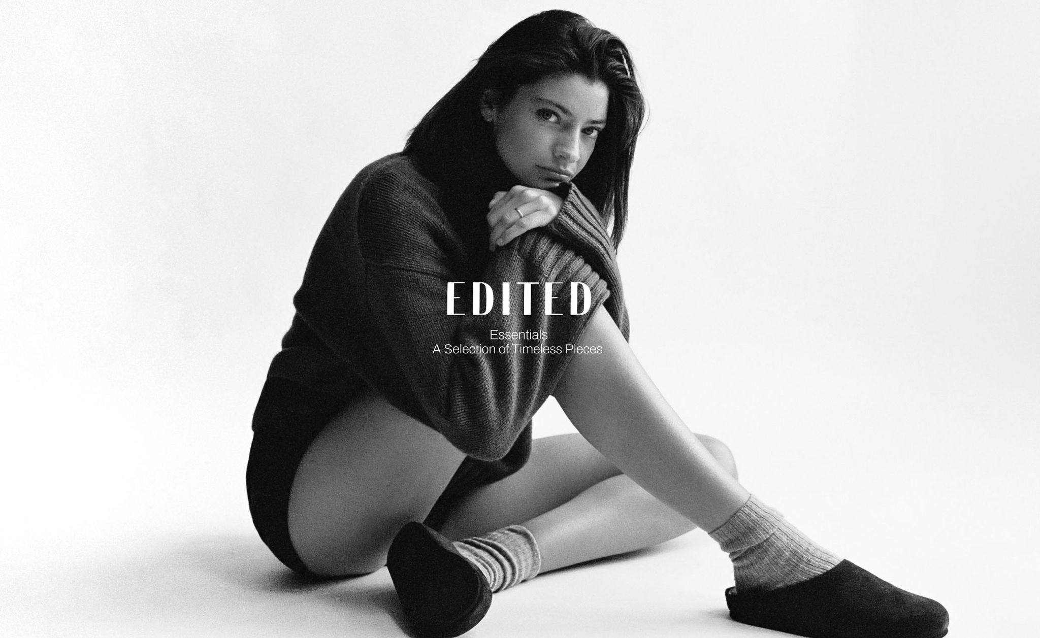 Black and white image of a woman sitting on the floor, wearing a sweater, shorts, socks, and slippers, with text that reads "EDITED Essentials."