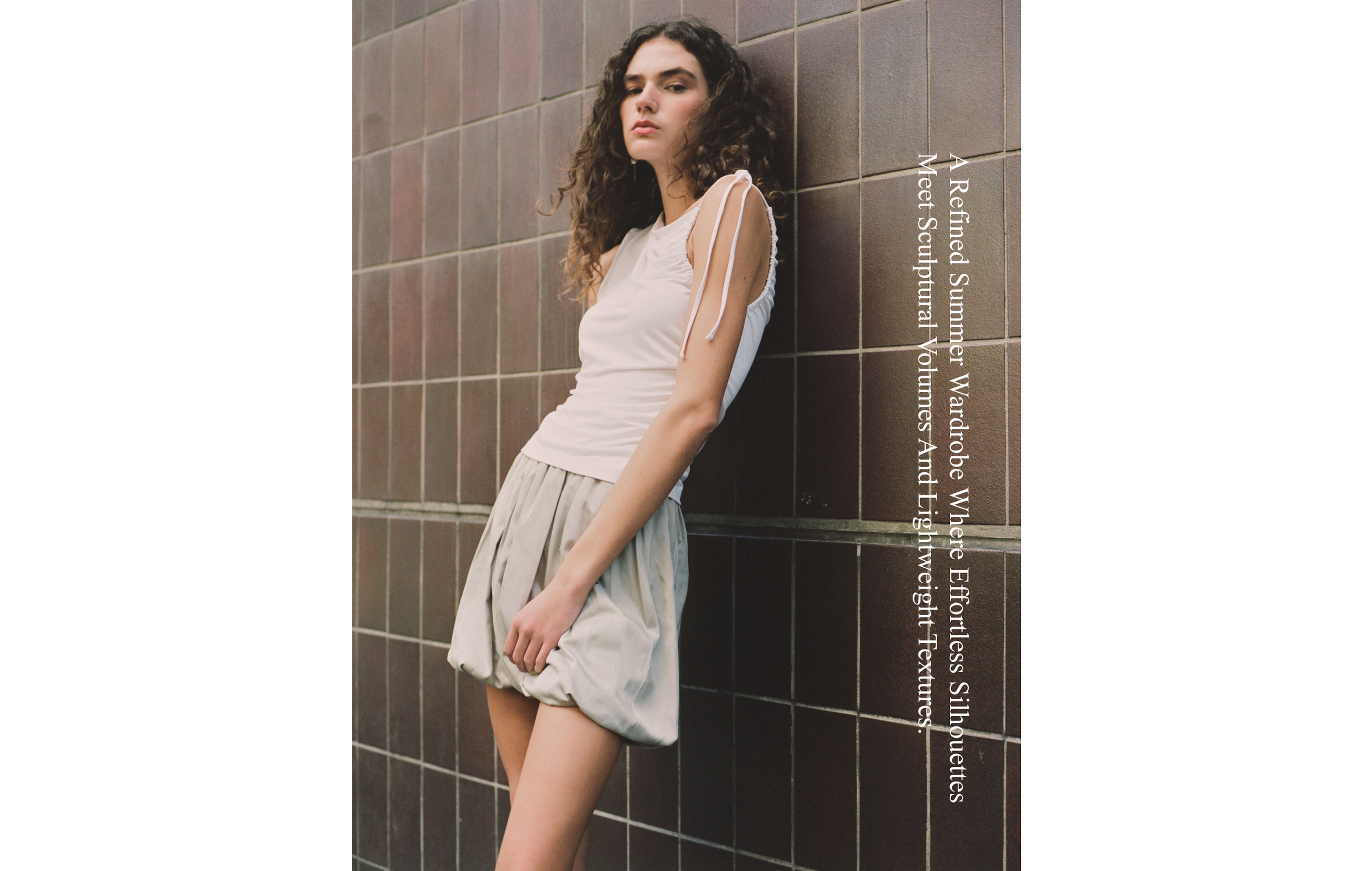 Young woman in a white sleeveless top and gray bubble skirt leaning against a tiled wall with text about a refined summer wardrobe.