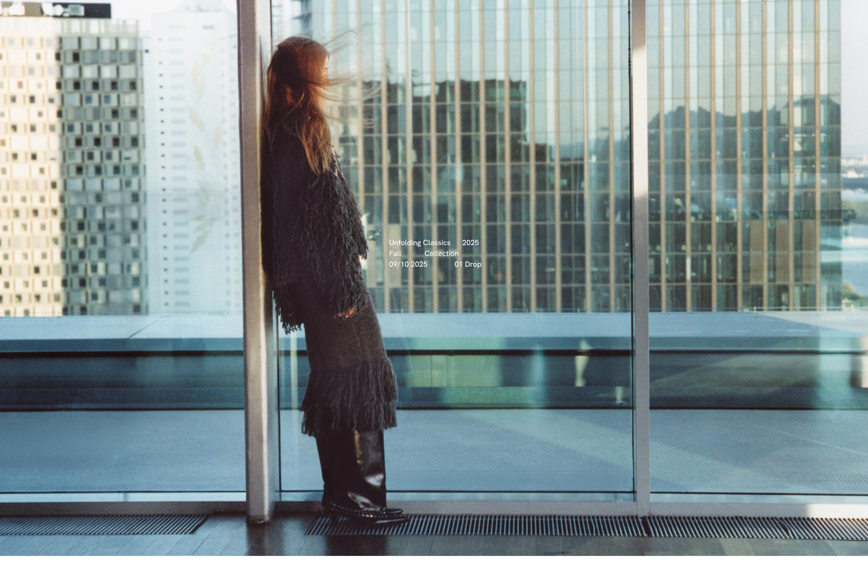 Woman in a fringed coat and boots leans against a glass wall, with a cityscape of tall buildings in the background.