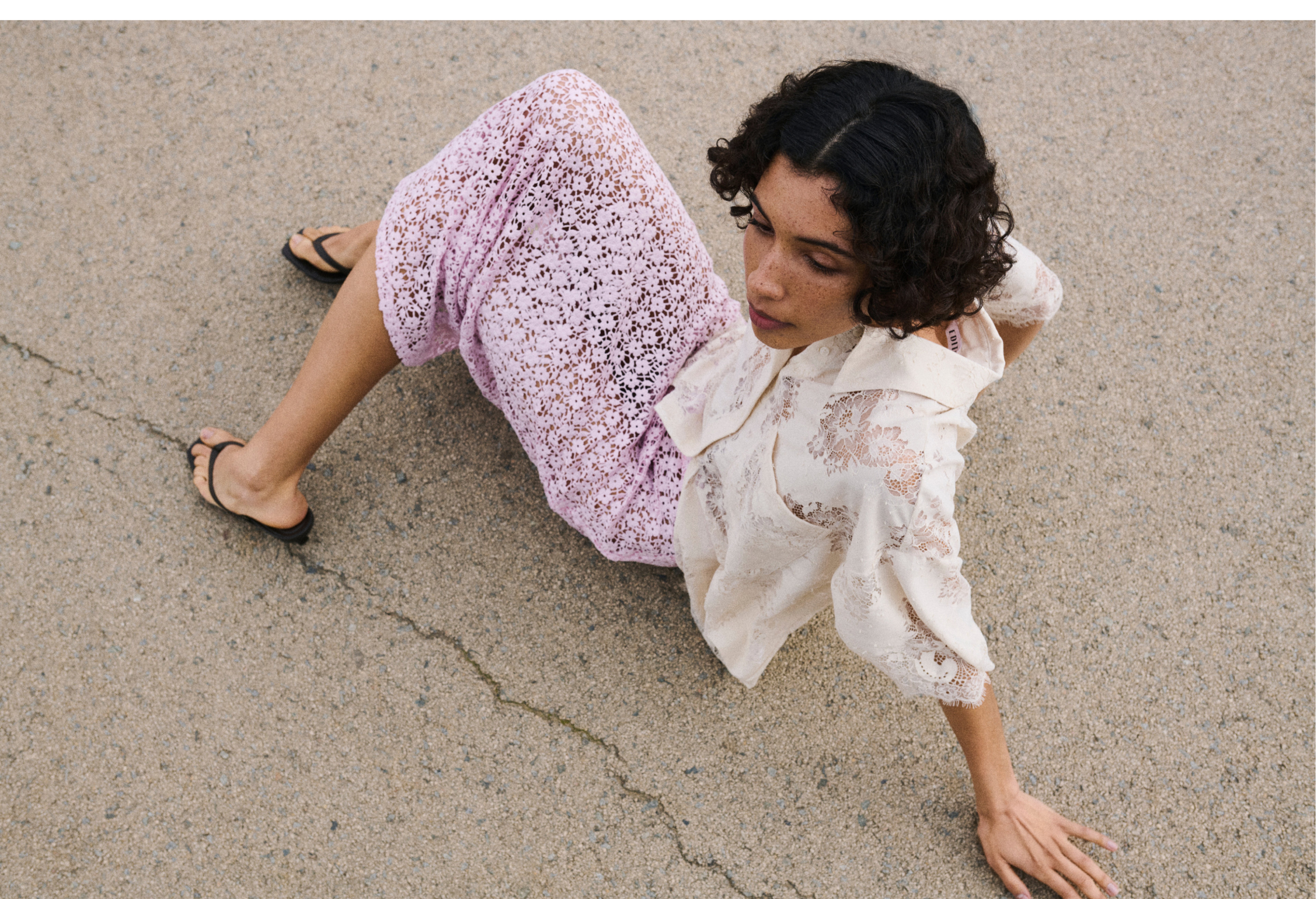 Person sitting on concrete, wearing a cream lace blouse and pink crochet pants, gazing to the side with one hand on the ground.