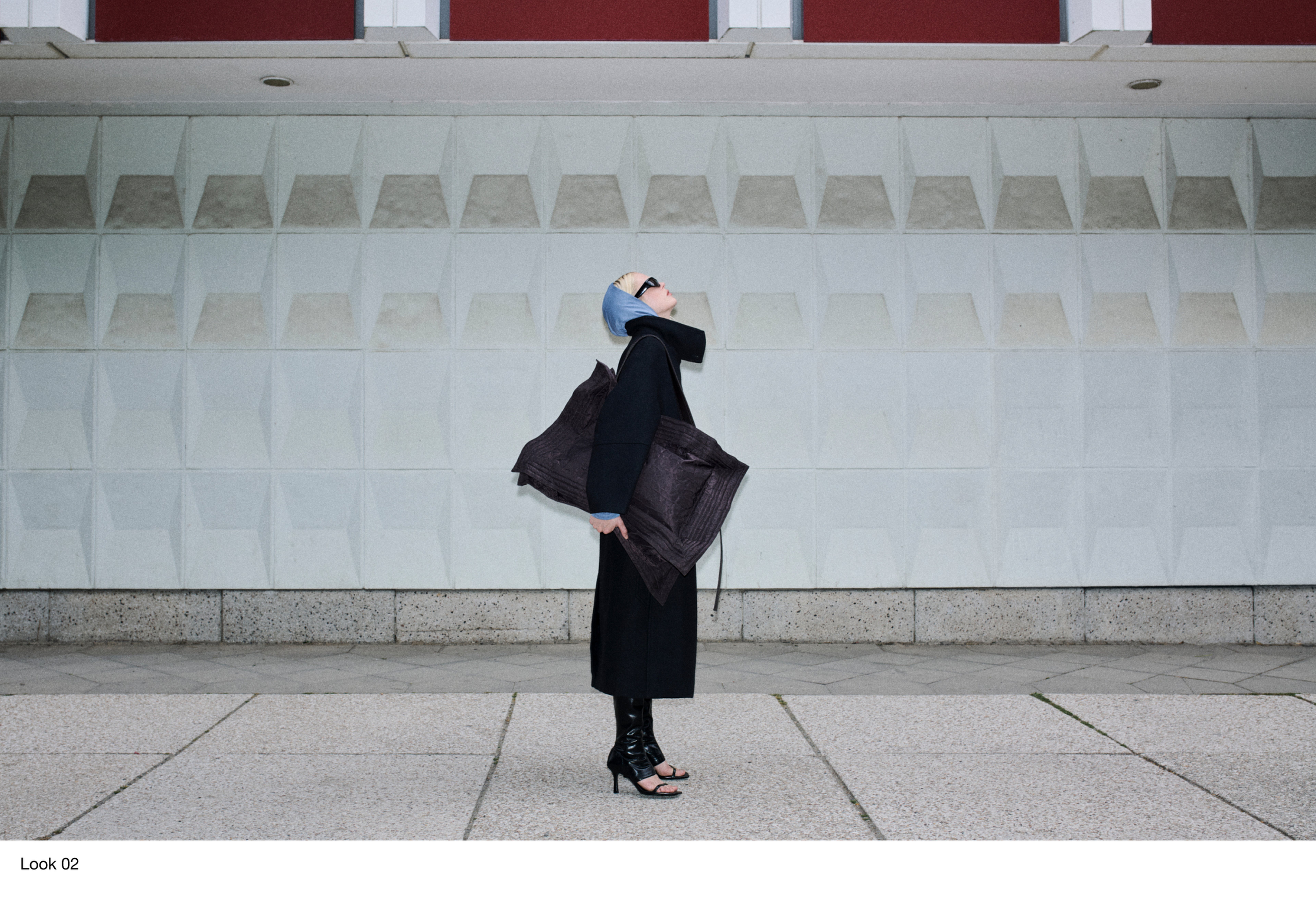 Person in a black coat, wearing sunglasses and a blue headscarf, holding a large textured bag, standing in front of a geometric wall.