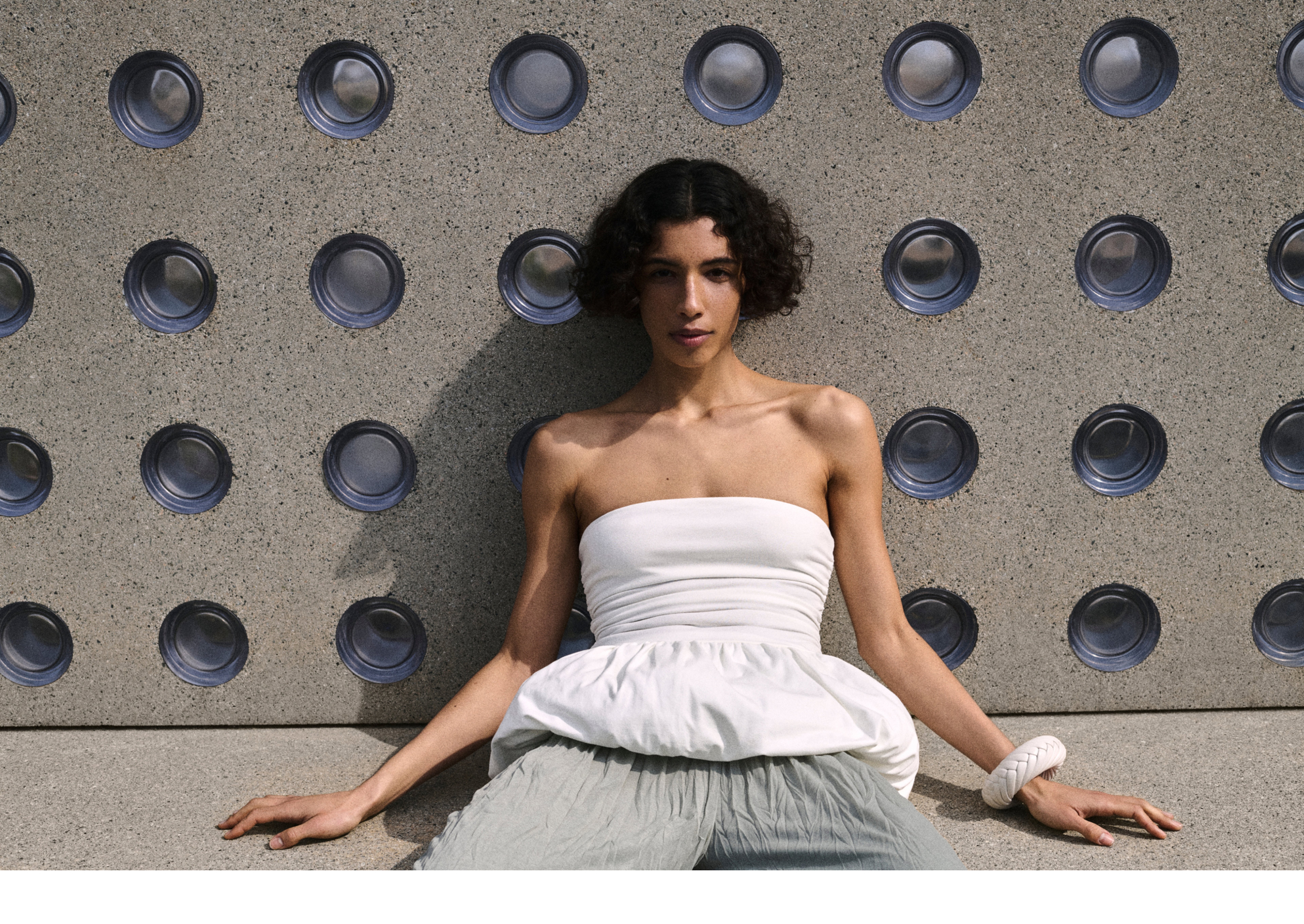 Person in a white strapless top and gray pants sits against a wall with circular patterns, casting a soft shadow.