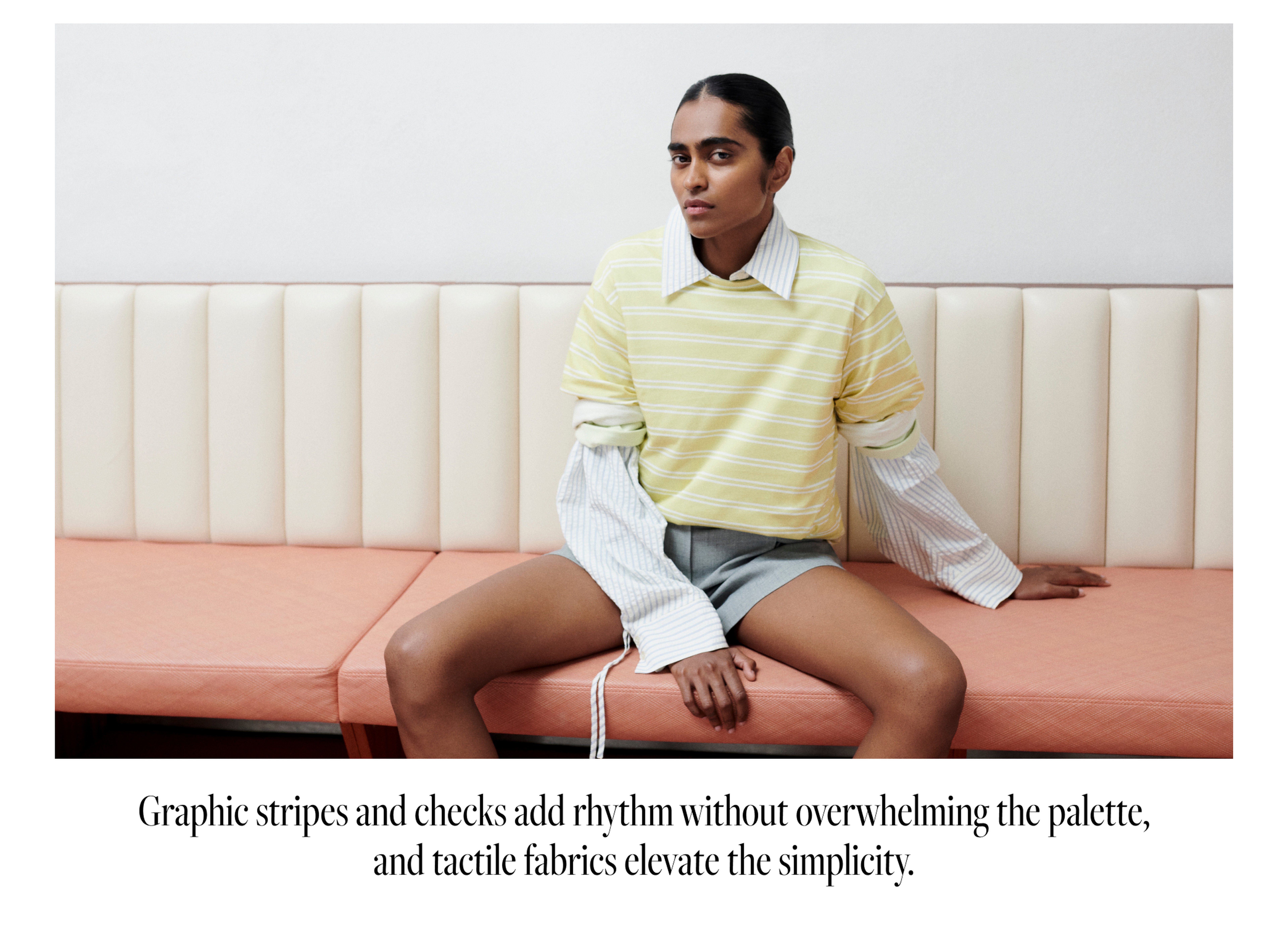 Person seated on a peach bench, wearing layered striped clothing, with neutral expression against a white wall.