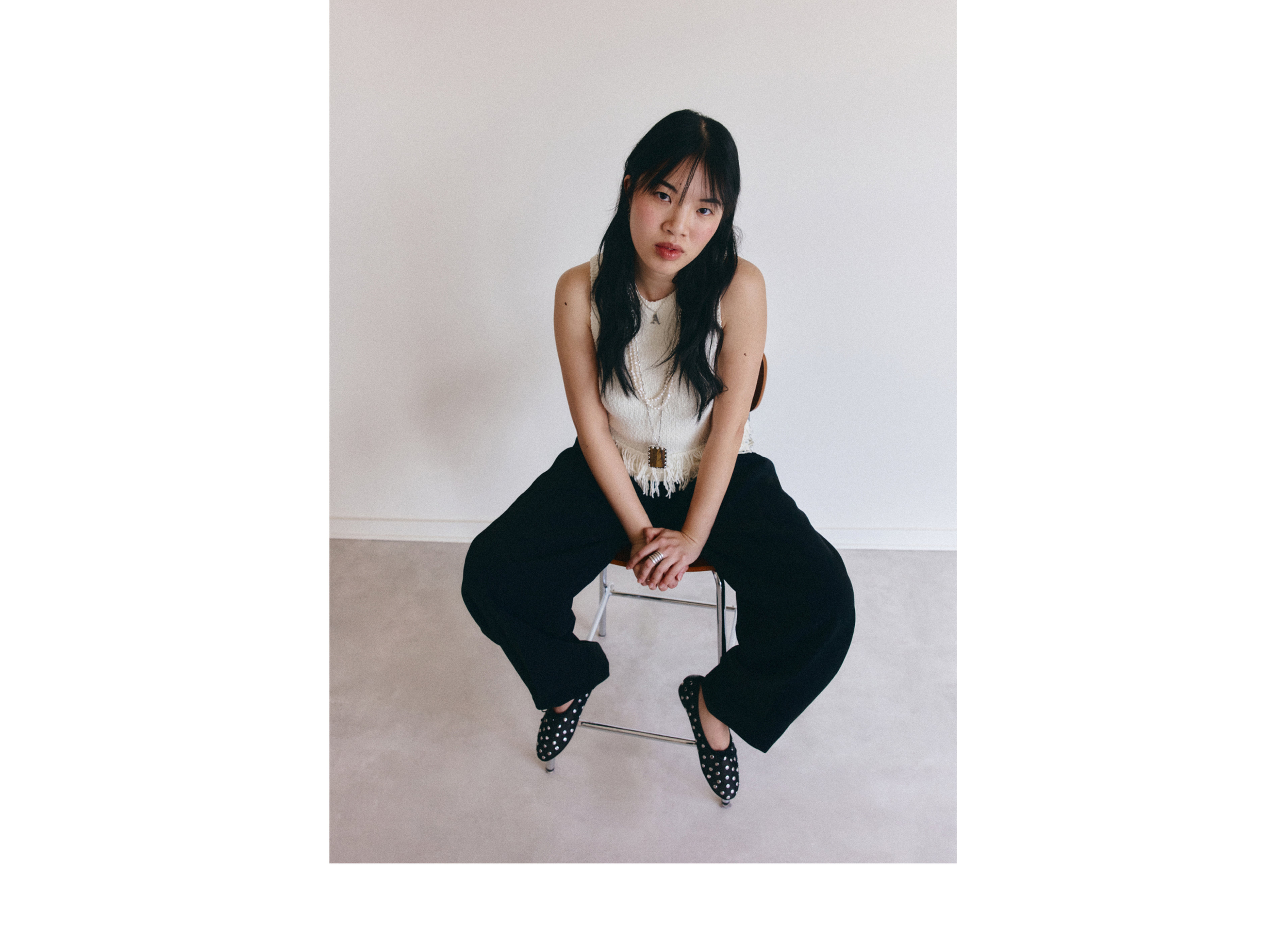 Woman in a white sleeveless top and black pants sits on a stool against a plain white wall, looking directly at the camera.