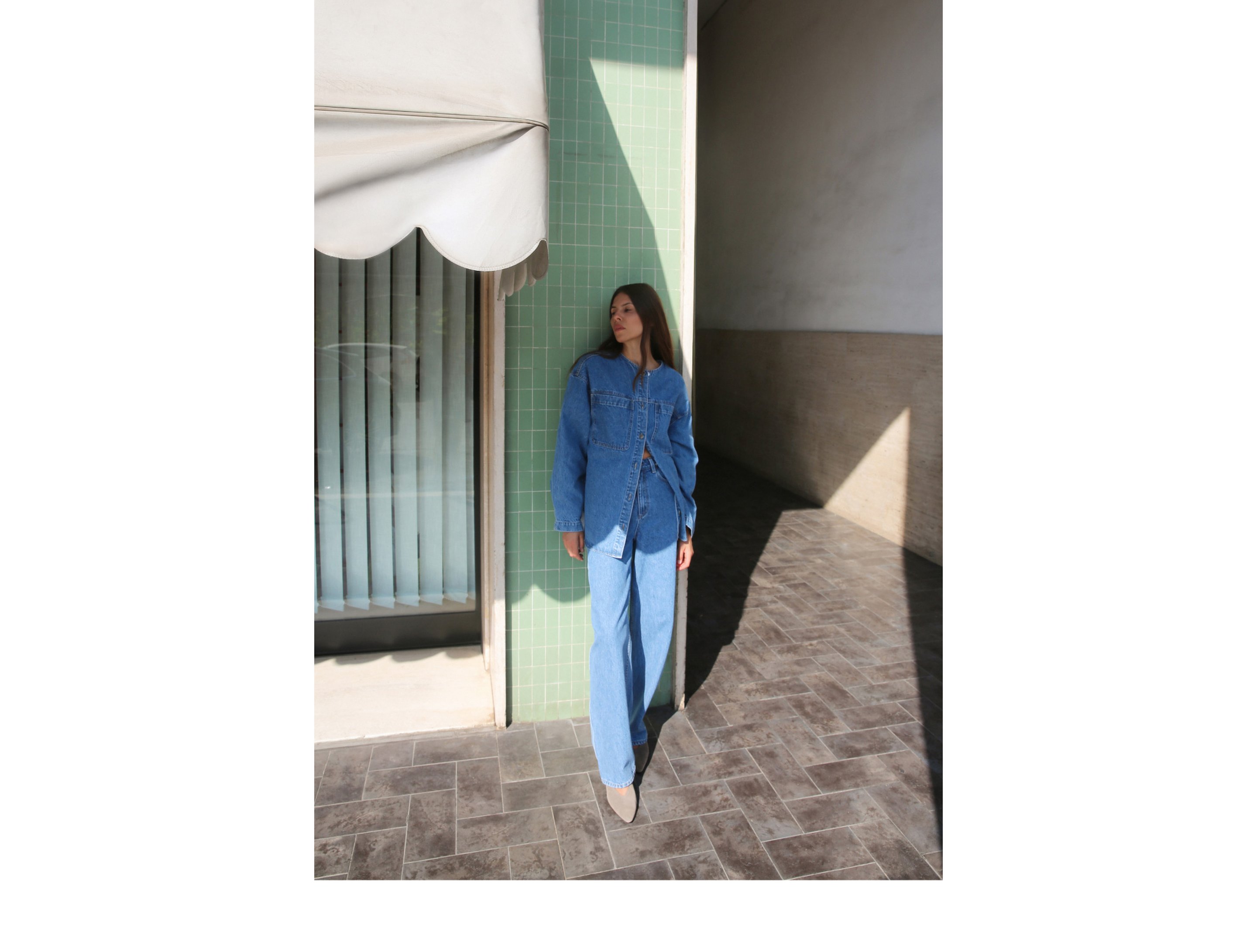 Woman in denim outfit stands against a green tiled wall, casting a shadow on the sunlit pavement.