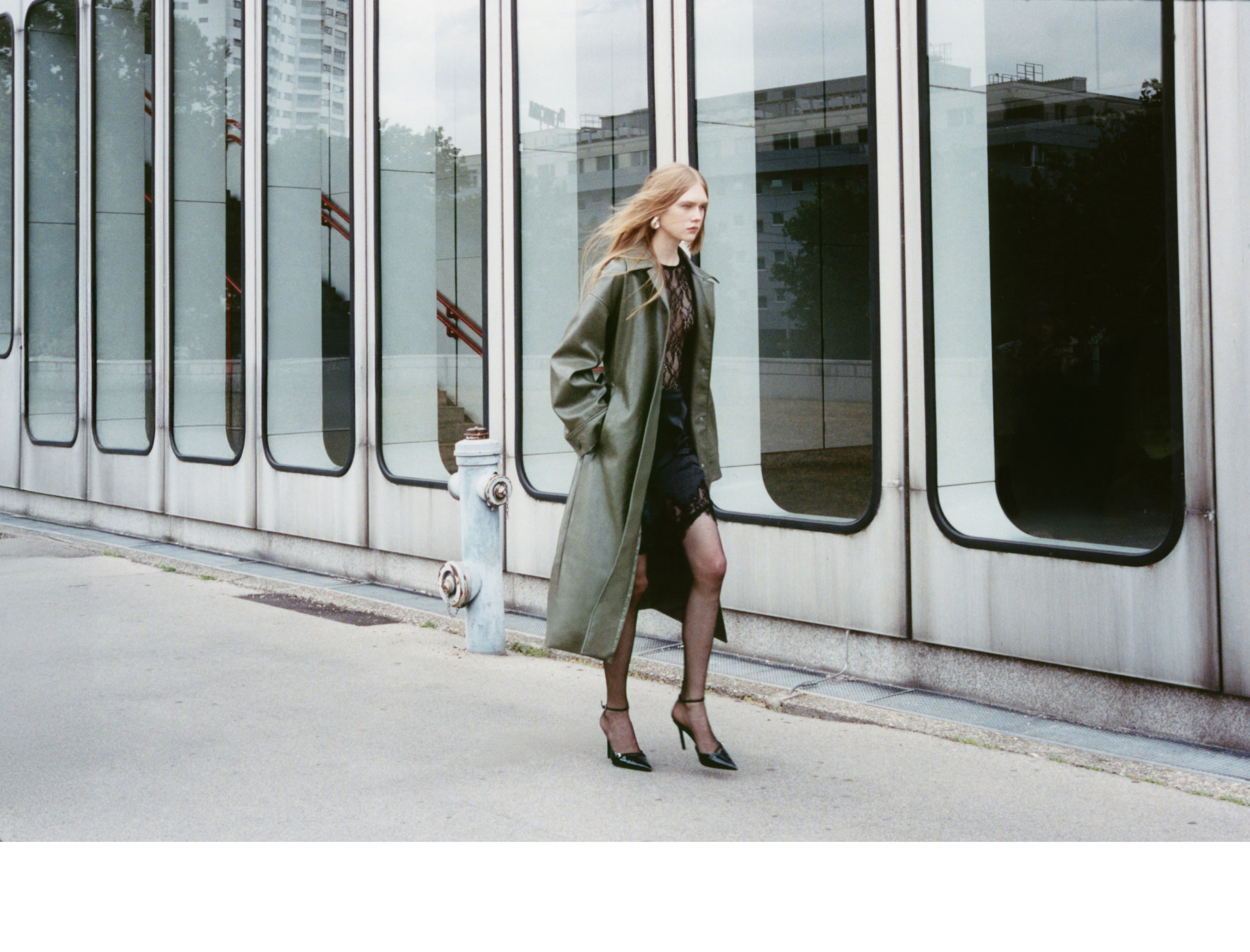 Person in a long green coat and heels walking past a modern building with large windows and a fire hydrant on the sidewalk.