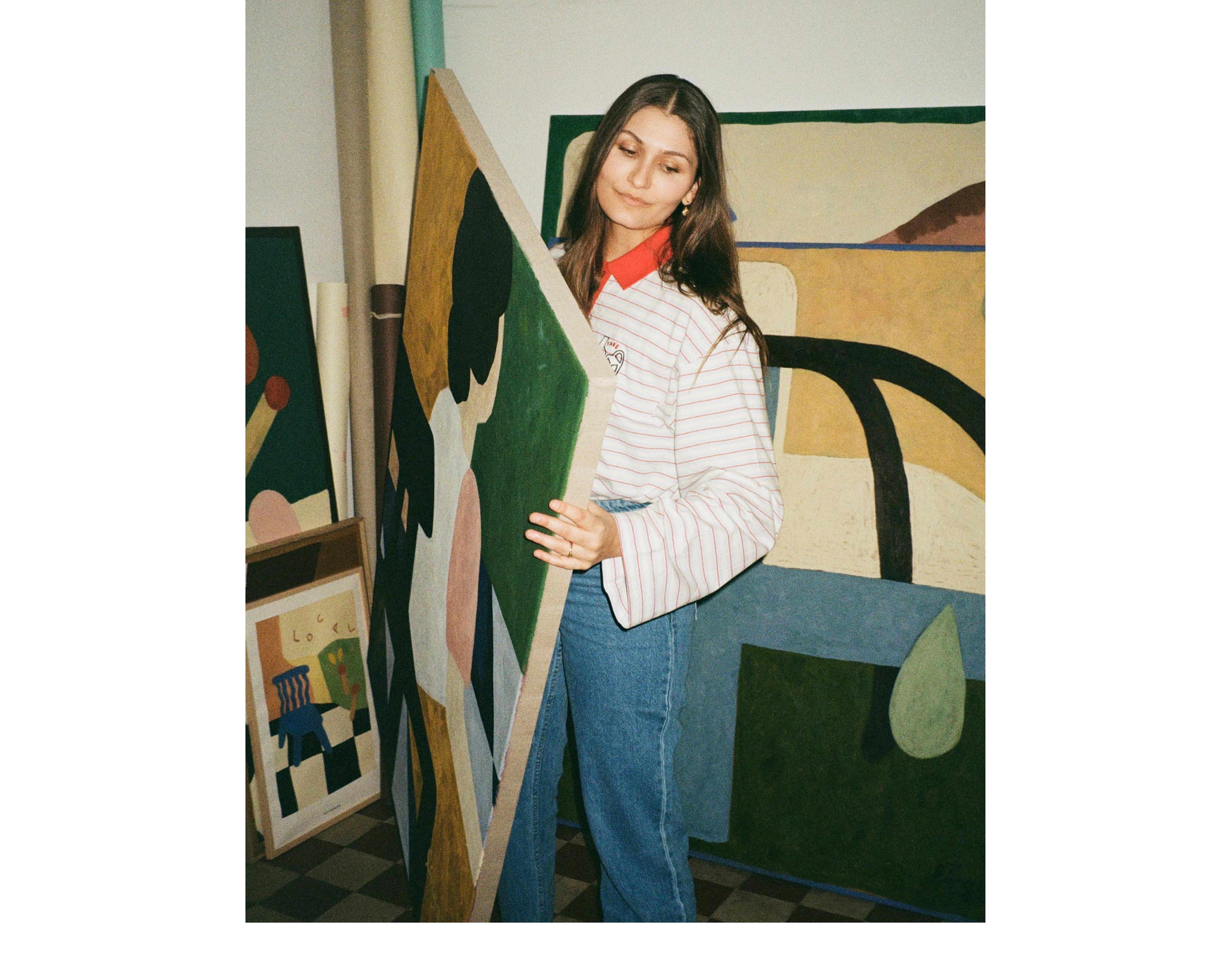 Woman in a striped shirt and jeans holding a large abstract painting in an art studio with multiple artworks around.