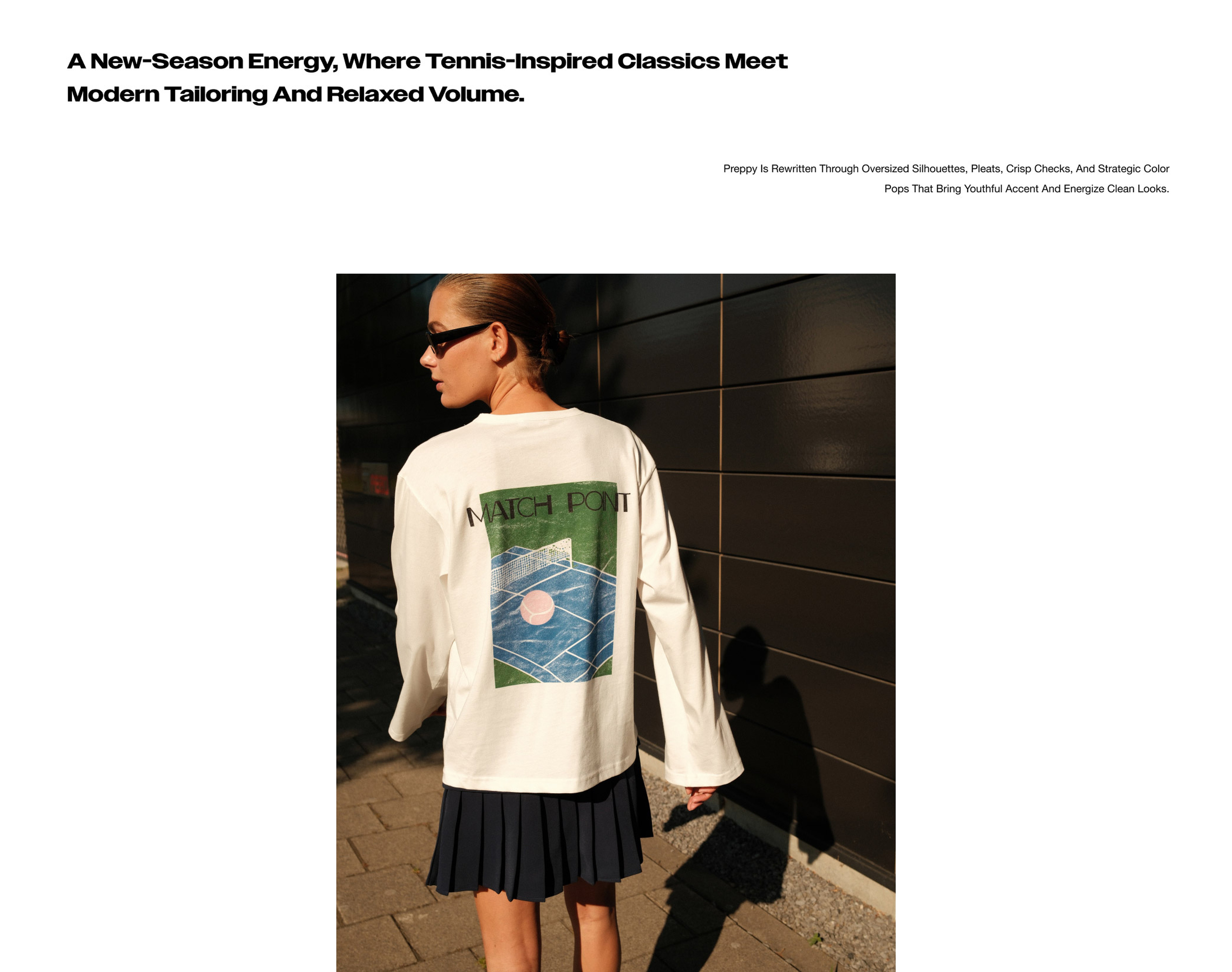 A person in a white shirt with a tennis court graphic, pleated skirt, and sunglasses walks along a sunlit, dark-paneled building.