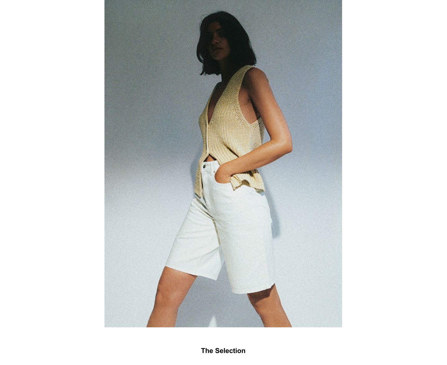 Woman in a sleeveless beige top and white shorts stands against a plain background, hands in pockets, with soft lighting creating shadows.