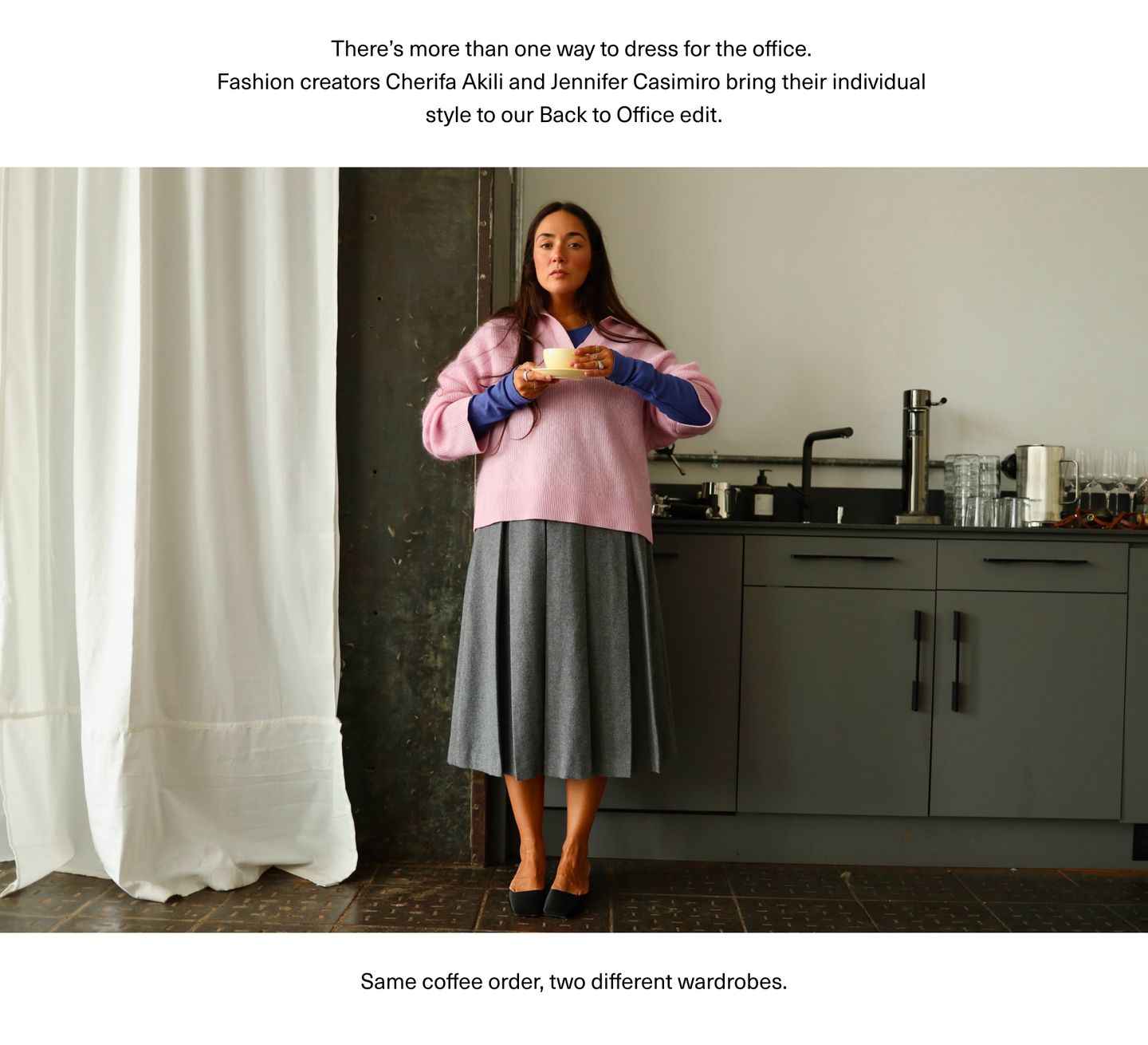 A woman wearing a pink sweater over a blue shirt and gray skirt stands in front of a kitchen counter, holding a coffee cup.