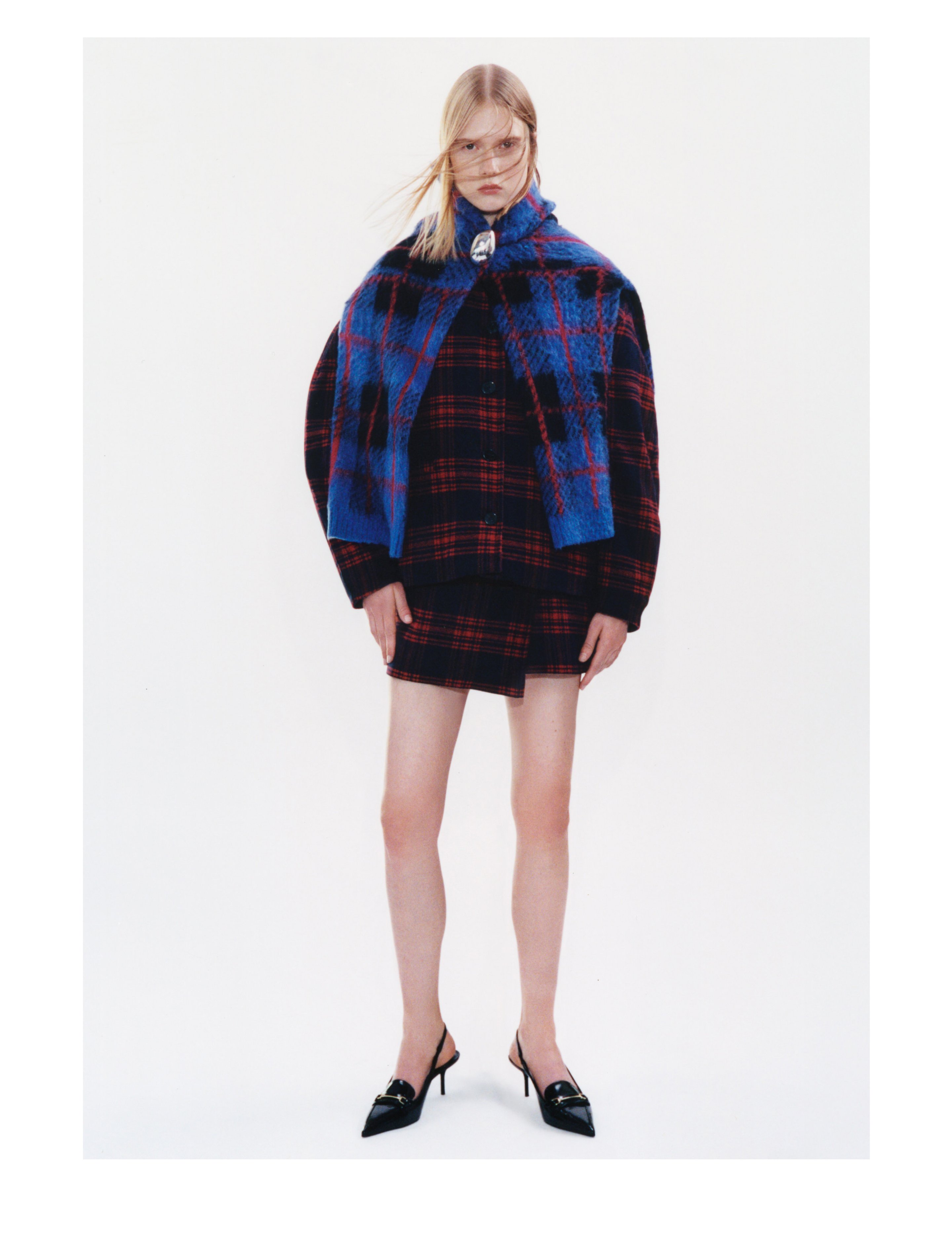 Person wearing a blue and red plaid outfit with a matching scarf, black pointed heels, and glasses, standing against a plain white background.
