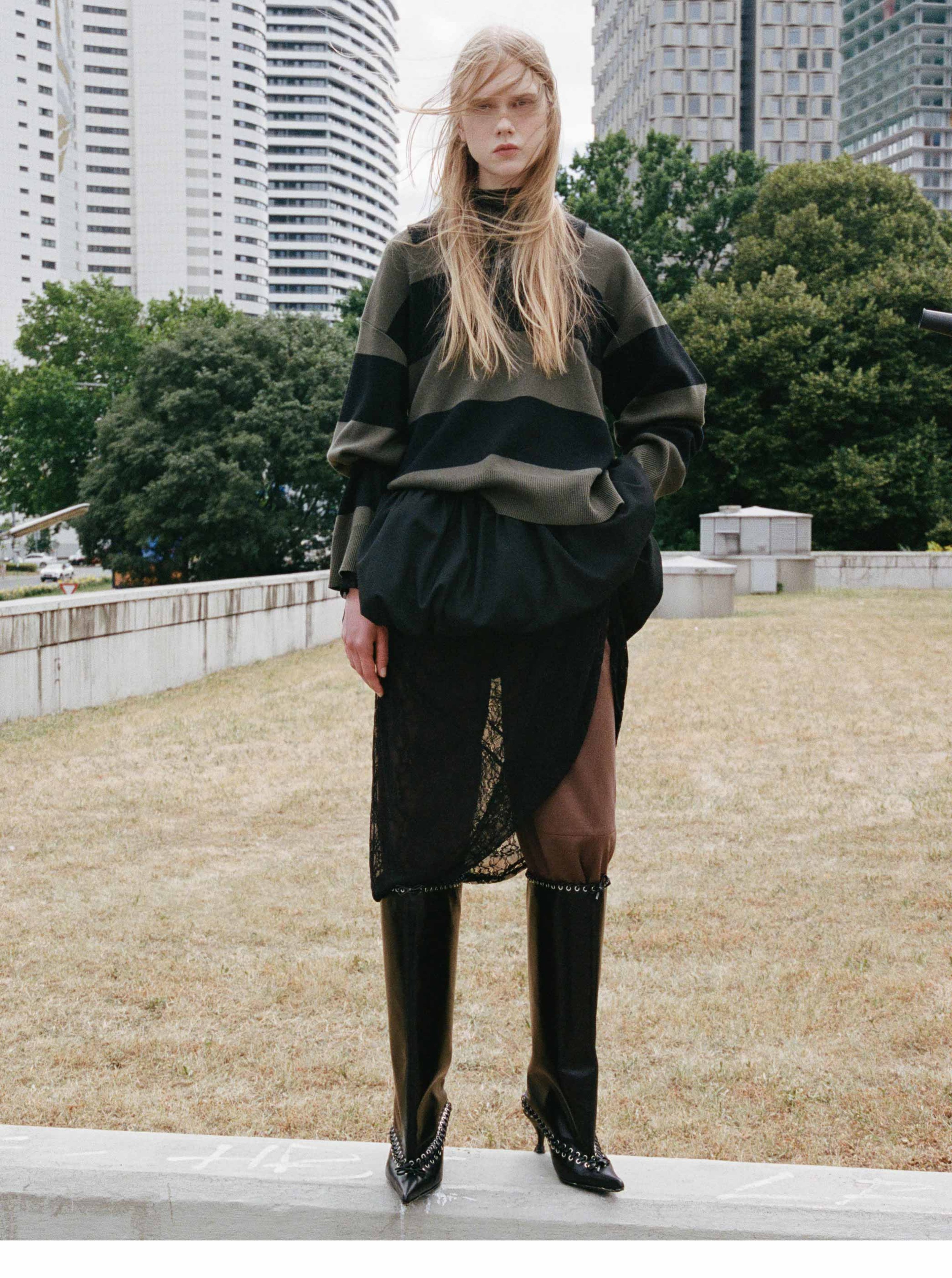A person stands outdoors wearing a green and black striped sweater, layered skirt, and tall black boots, with buildings and trees in the background.