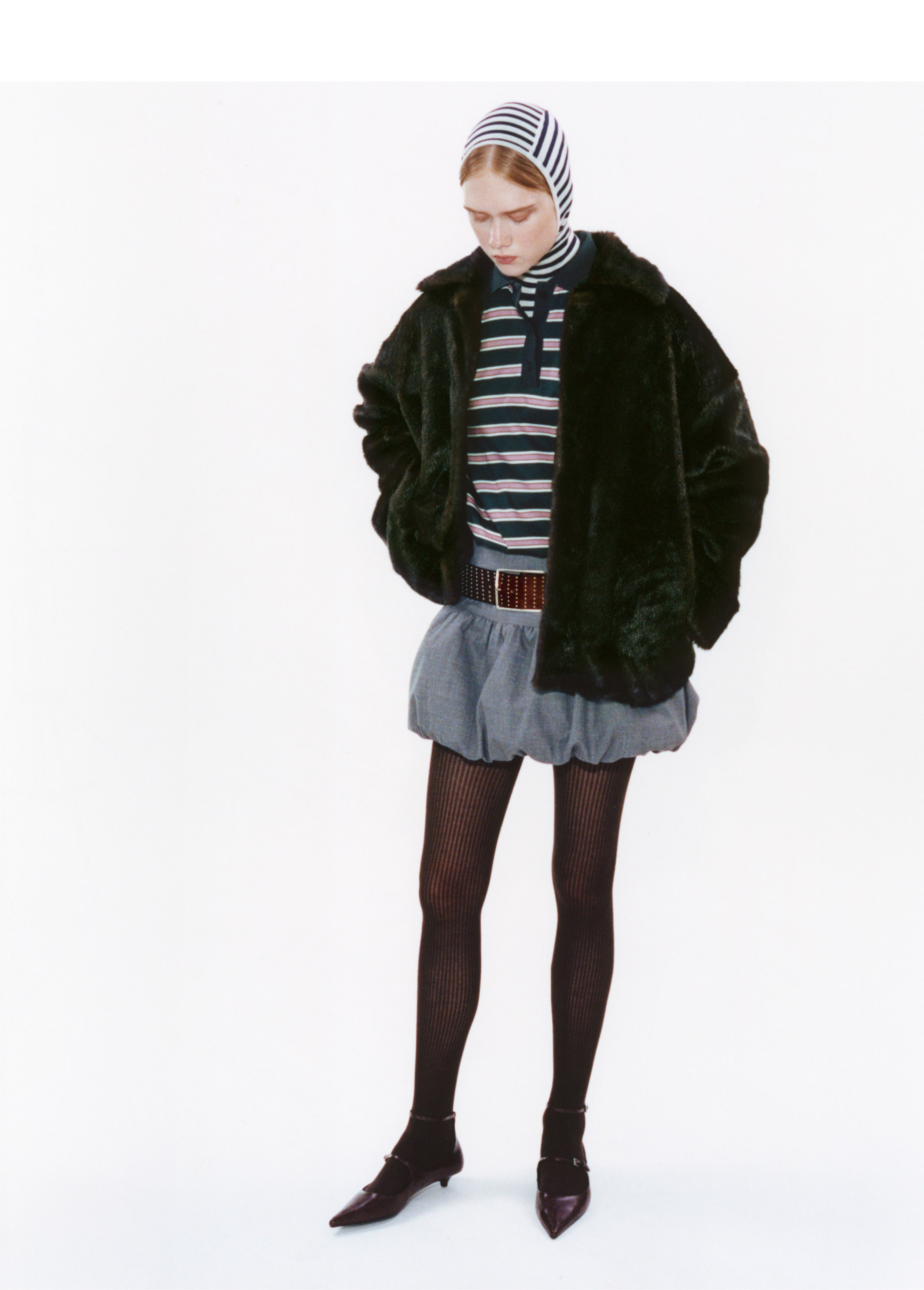 Person in a striped headscarf, dark fur coat, striped shirt, short skirt, and patterned tights, standing on a white background.