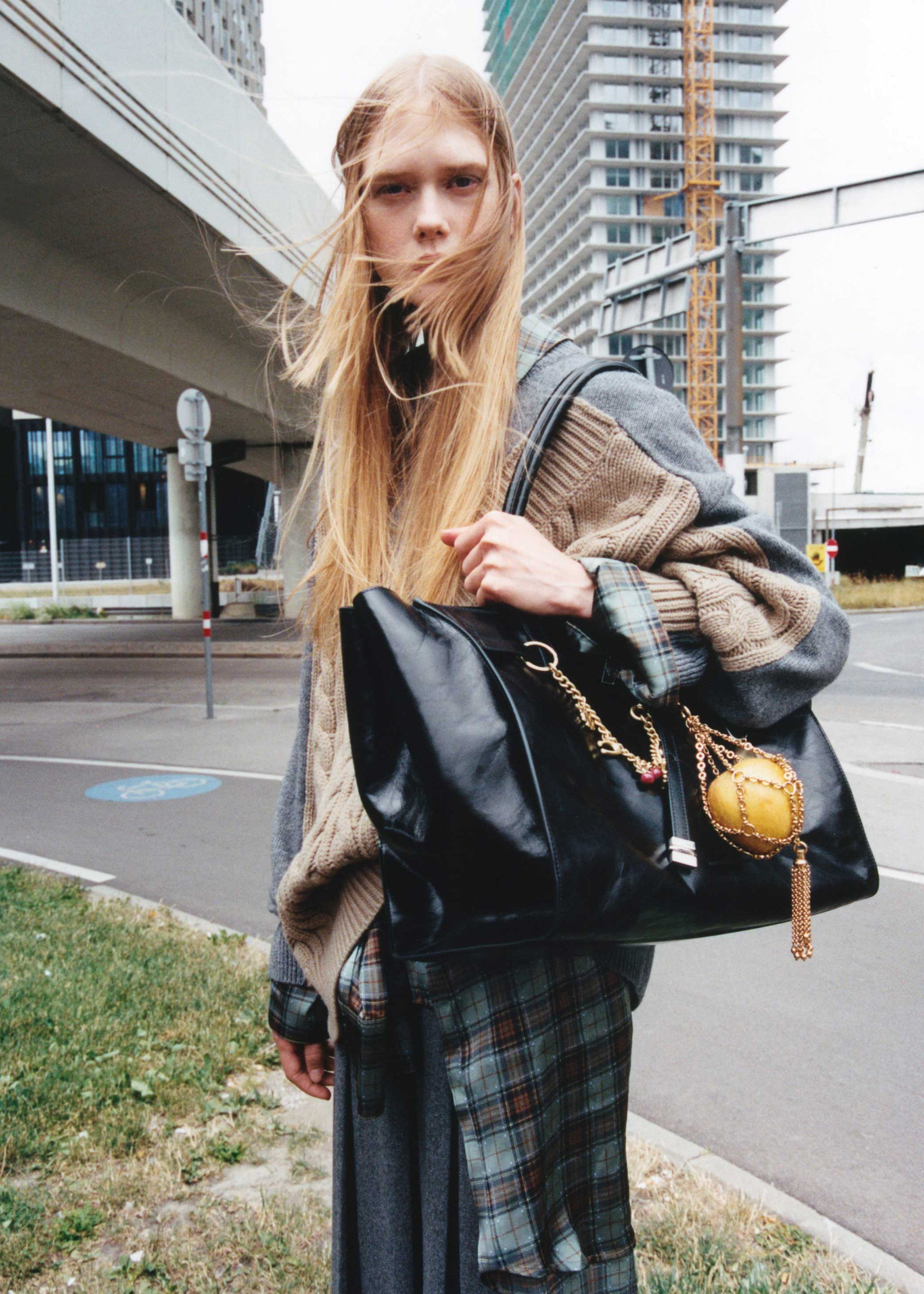 Person with long hair in layered clothing holds a large black bag, standing outdoors near a road and modern buildings.