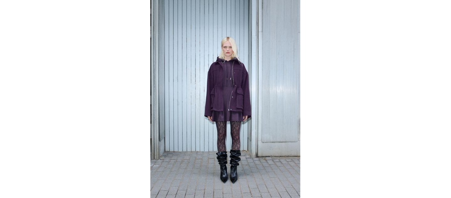 A person stands against a light gray corrugated metal wall, wearing a purple oversized jacket, lace tights, and black slouched boots.