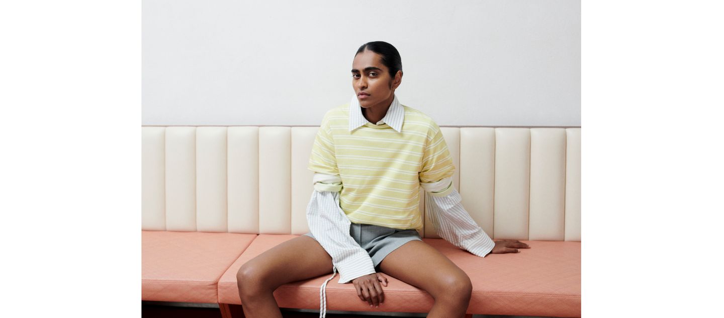 Person wearing a yellow striped shirt and gray shorts, sitting on a cushioned bench, looking thoughtfully to the side.