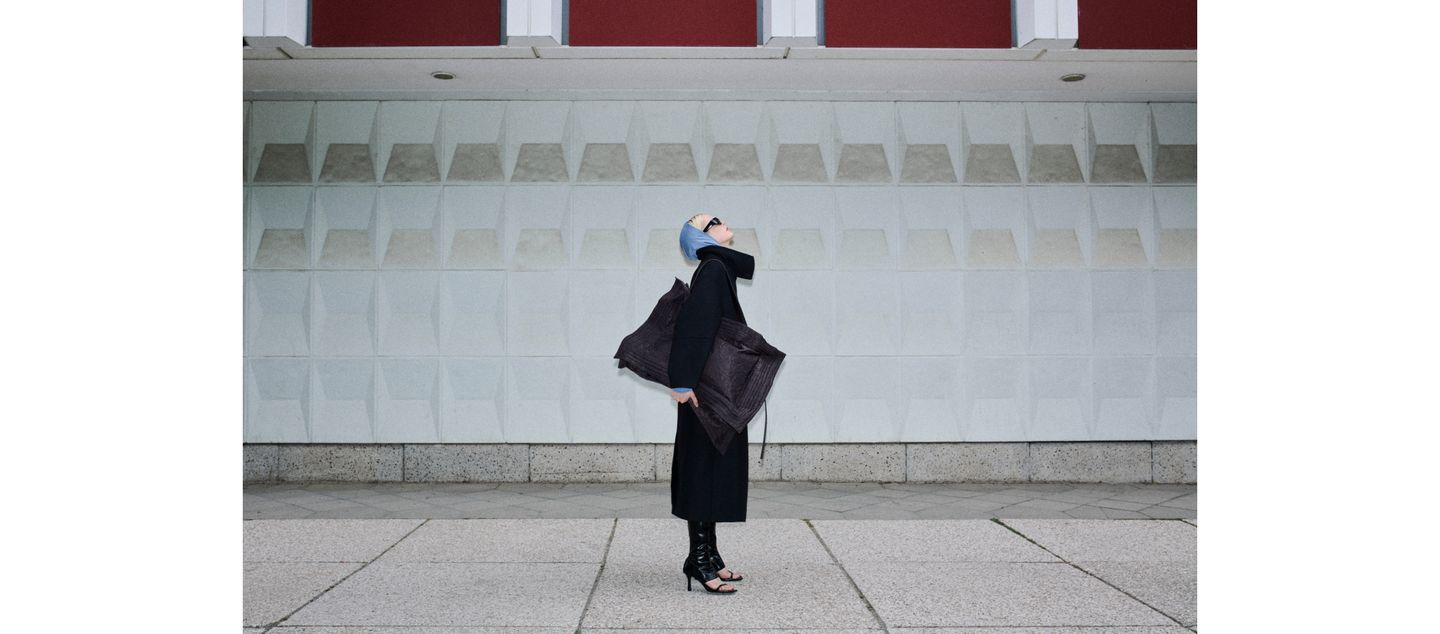 A person in a stylish black outfit and sunglasses stands against a geometric concrete wall, holding a large brown bag and looking upward.