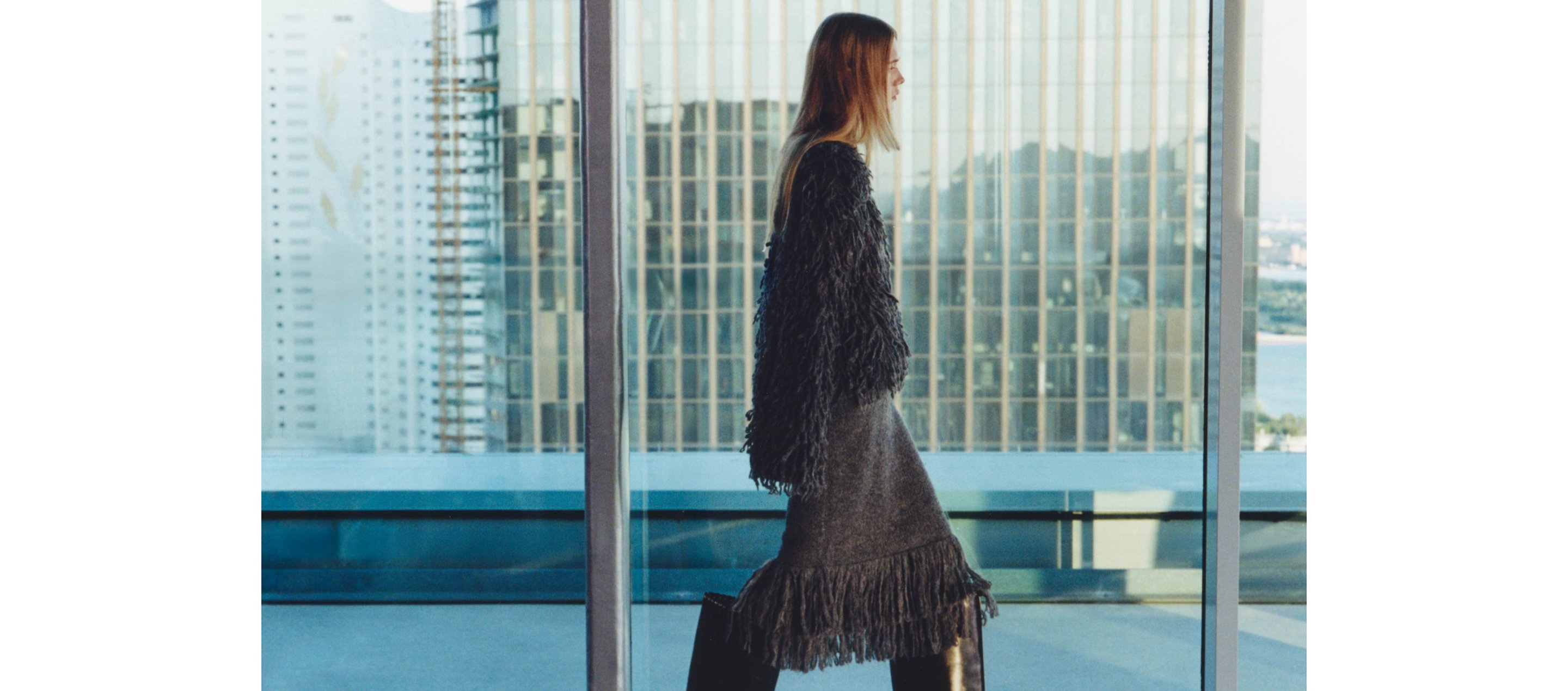 A woman in a fringed outfit and boots walks past large windows with a cityscape view in the background.