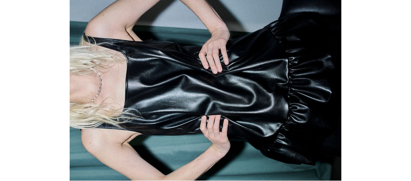 Person in a black, glossy leather top with a ruffled bottom, hands on hips, with long blond hair partially visible.