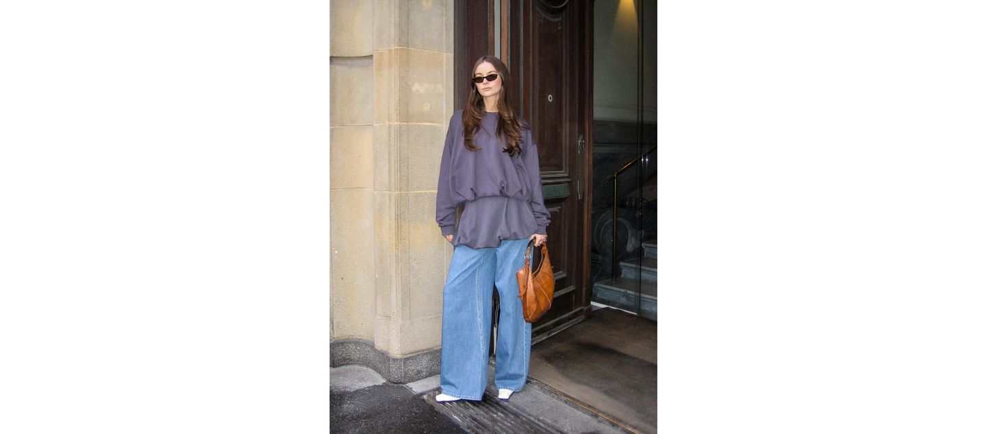 Woman in oversized shirt and wide jeans, wearing sunglasses, holds a brown bag while standing by a stone building entrance.