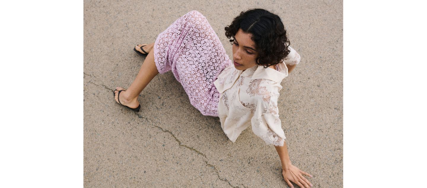 A woman with curly hair sits on a textured surface, wearing a pink lace skirt, a patterned blouse, and black sandals, viewed from above.