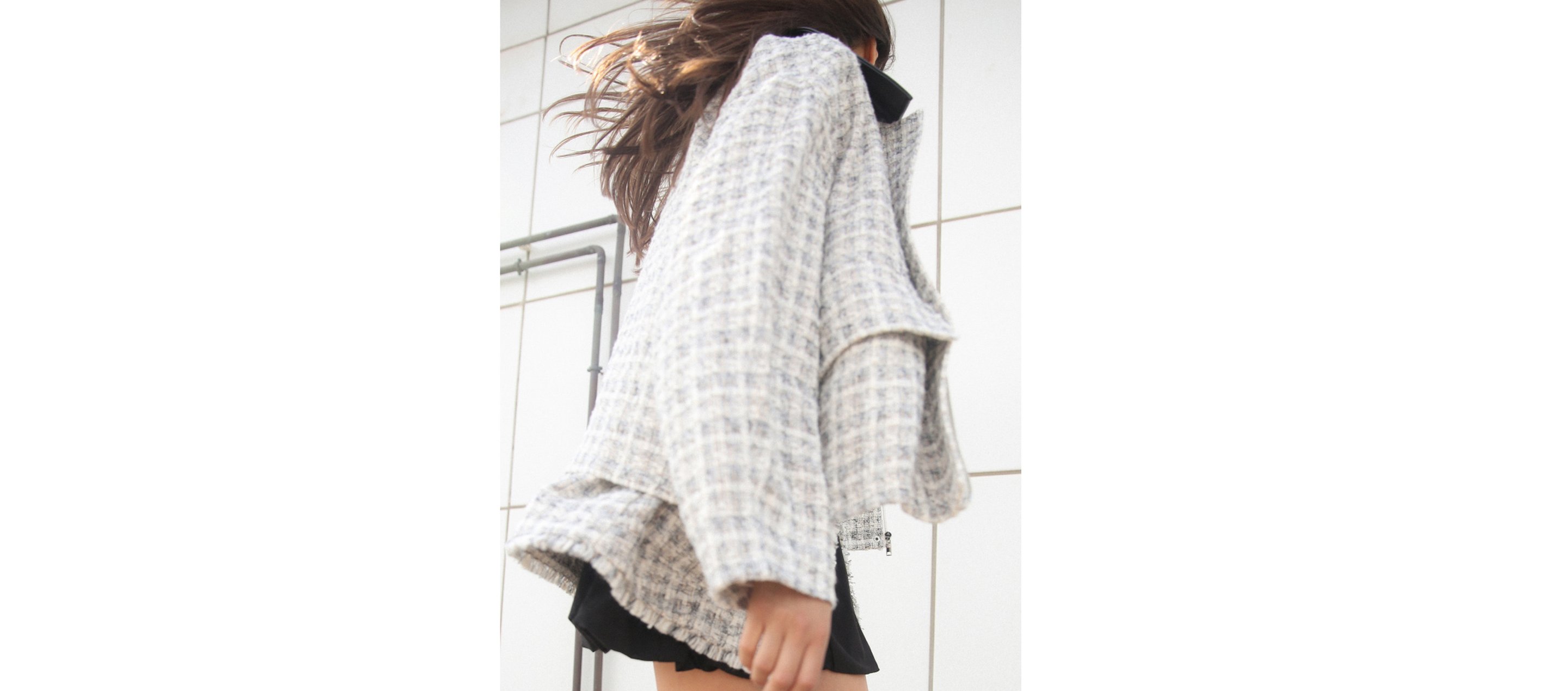 Person wearing a textured, oversized jacket and black skirt, with hair blowing in the wind, standing against a tiled wall.