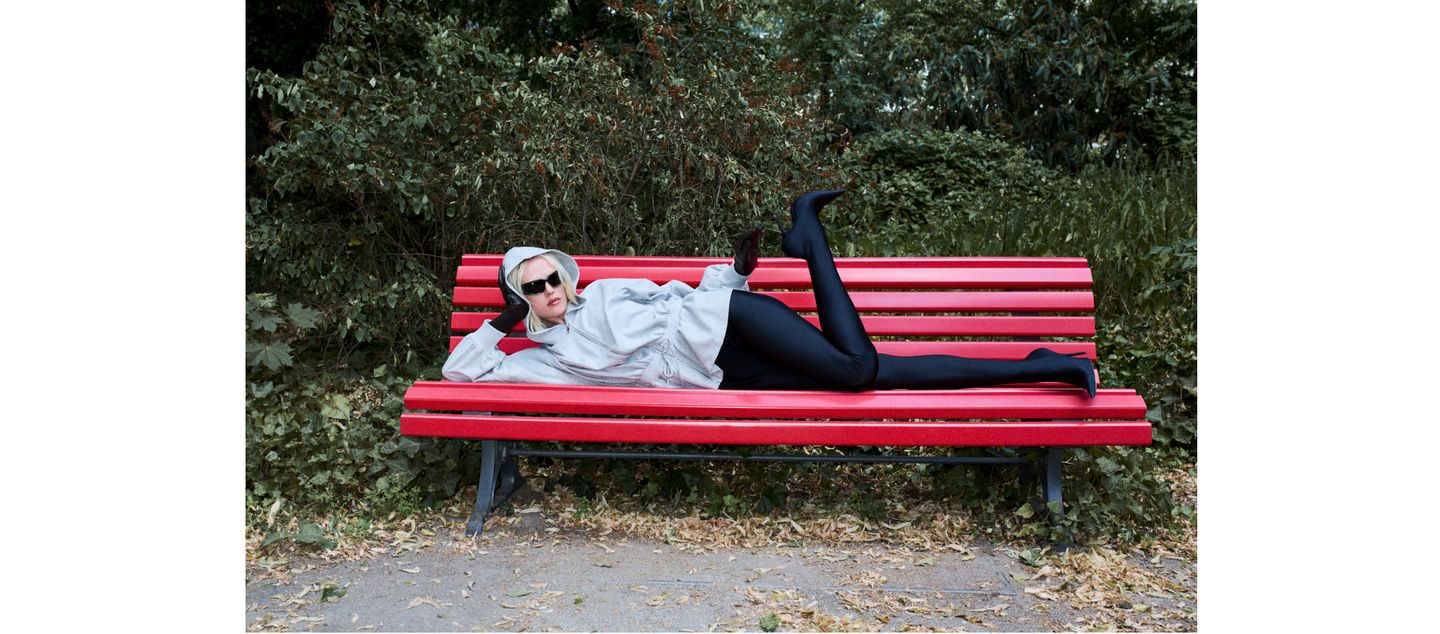 Person in a hoodie and sunglasses, posing on a red park bench in front of green foliage.