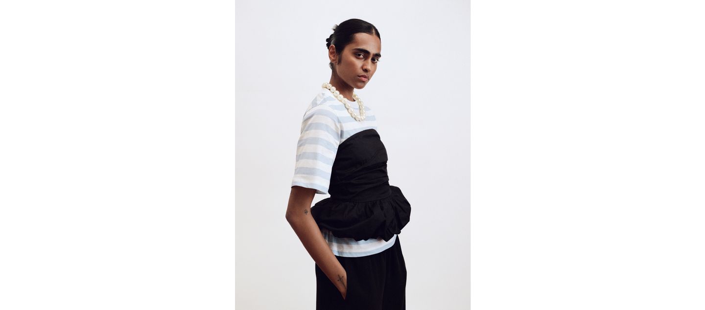 Person in a striped shirt and black peplum top, wearing a pearl necklace, standing against a plain white background.