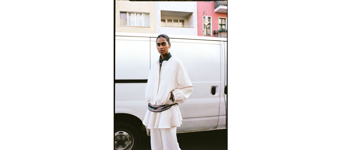 Person in a white, layered outfit stands on a street, posing in front of a white van, with colorful buildings in the background.
