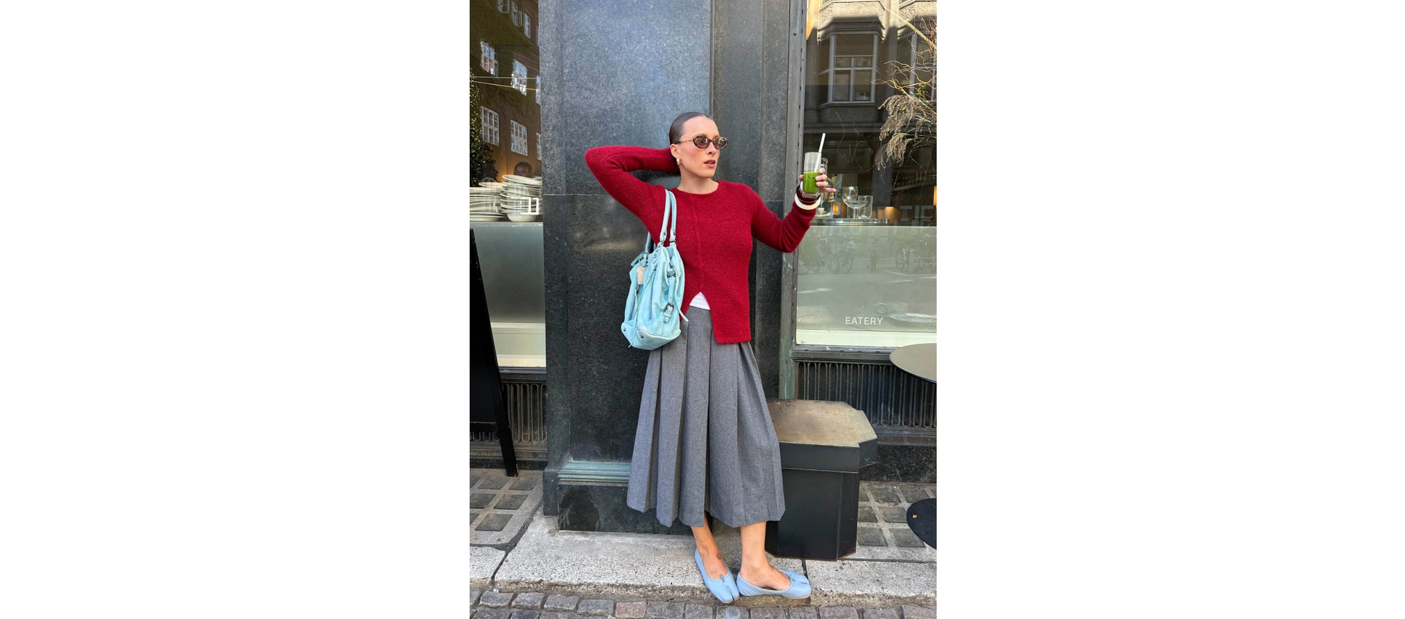 Person in a red sweater and gray skirt holds a drink and a phone, standing by a building with a blue bag and wearing sunglasses.